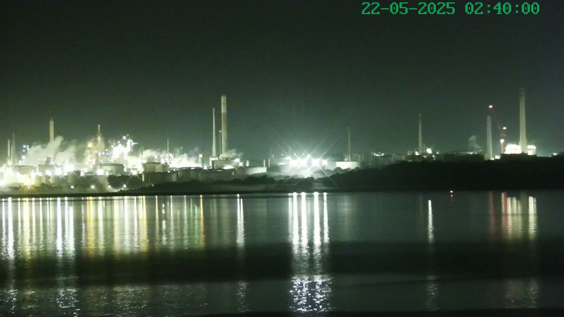A brightly lit industrial complex is reflected in a calm body of water at night.