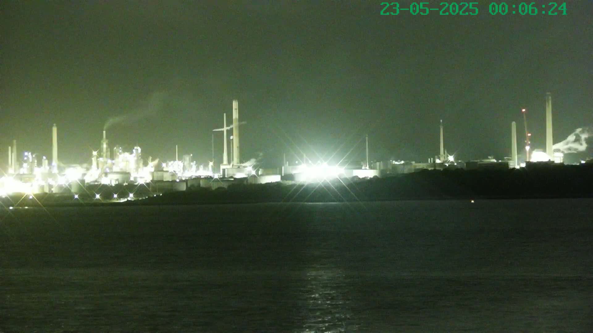 A brightly lit industrial plant at night is seen across a dark body of water under a dark sky.