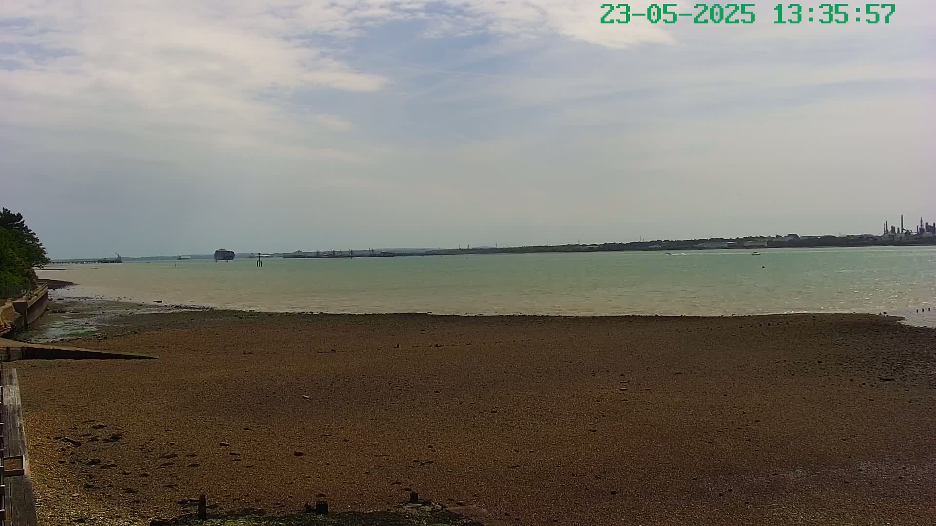 A mostly sunny day reveals a broad expanse of exposed mudflats leading to a calm, light-green body of water with a distant industrial shoreline.