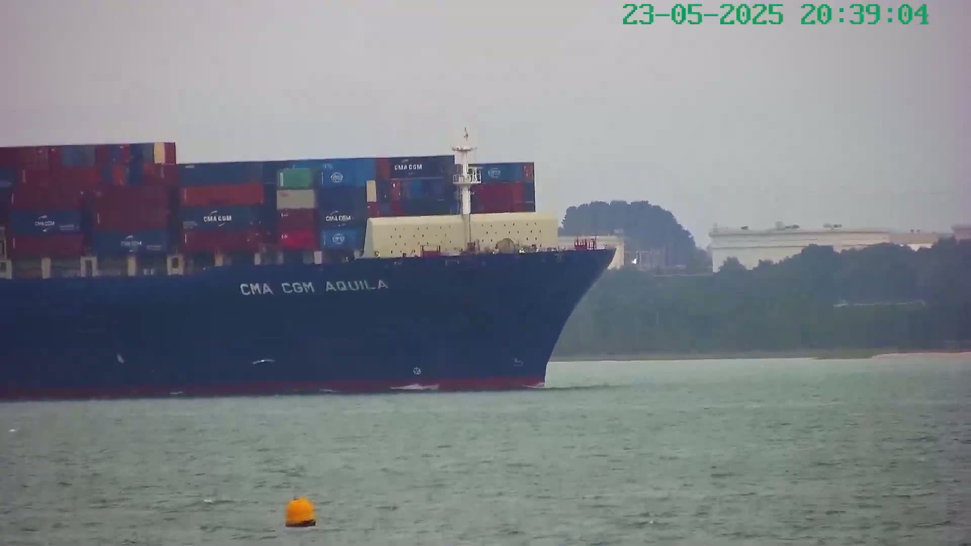 A large container ship, the CMA CGM Aquila, sails on a calm, overcast body of water past a hazy shoreline.