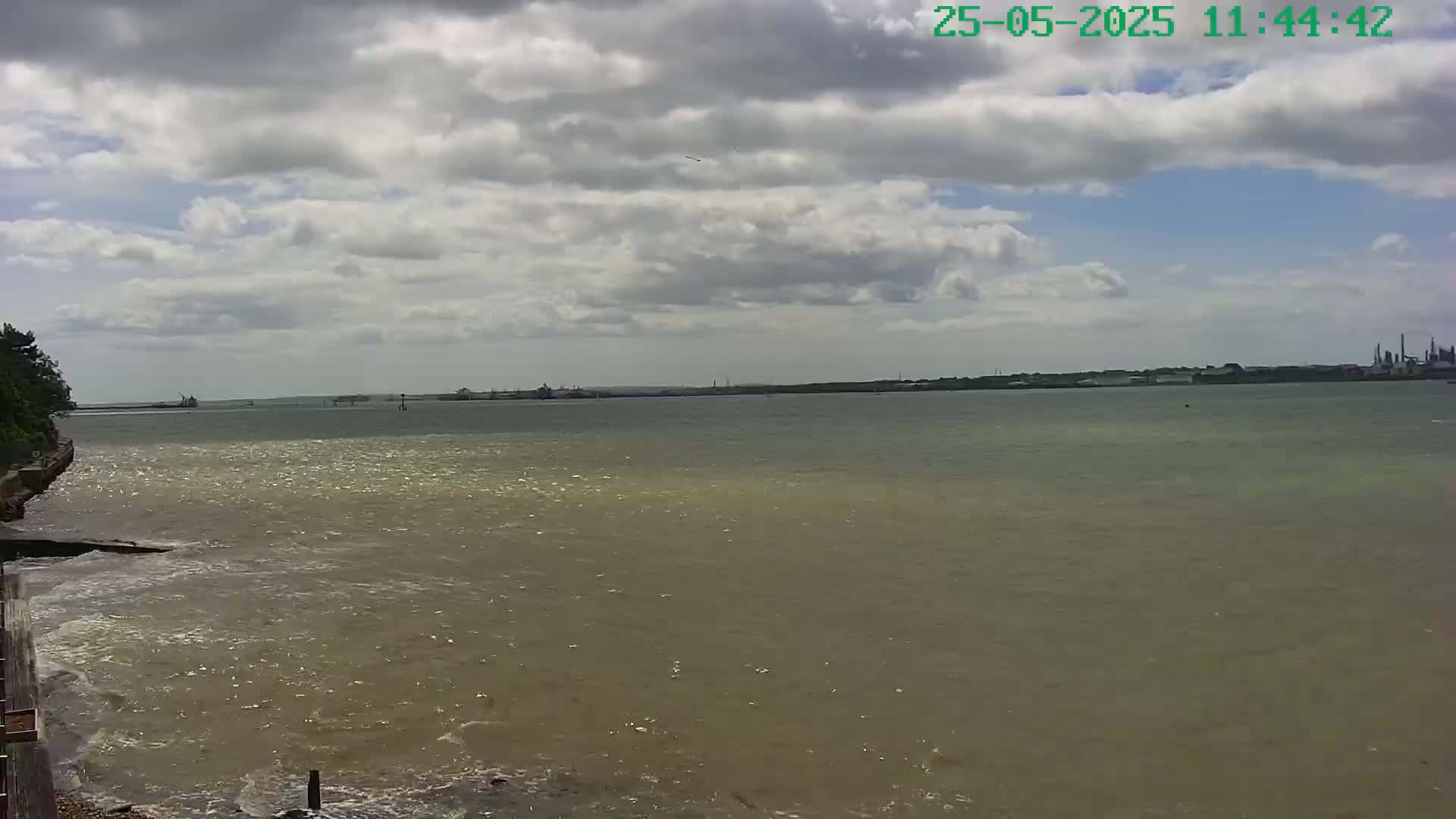 A partly cloudy day shows a view of a wide body of water, with a shoreline visible in the foreground and industrial structures in the distance.