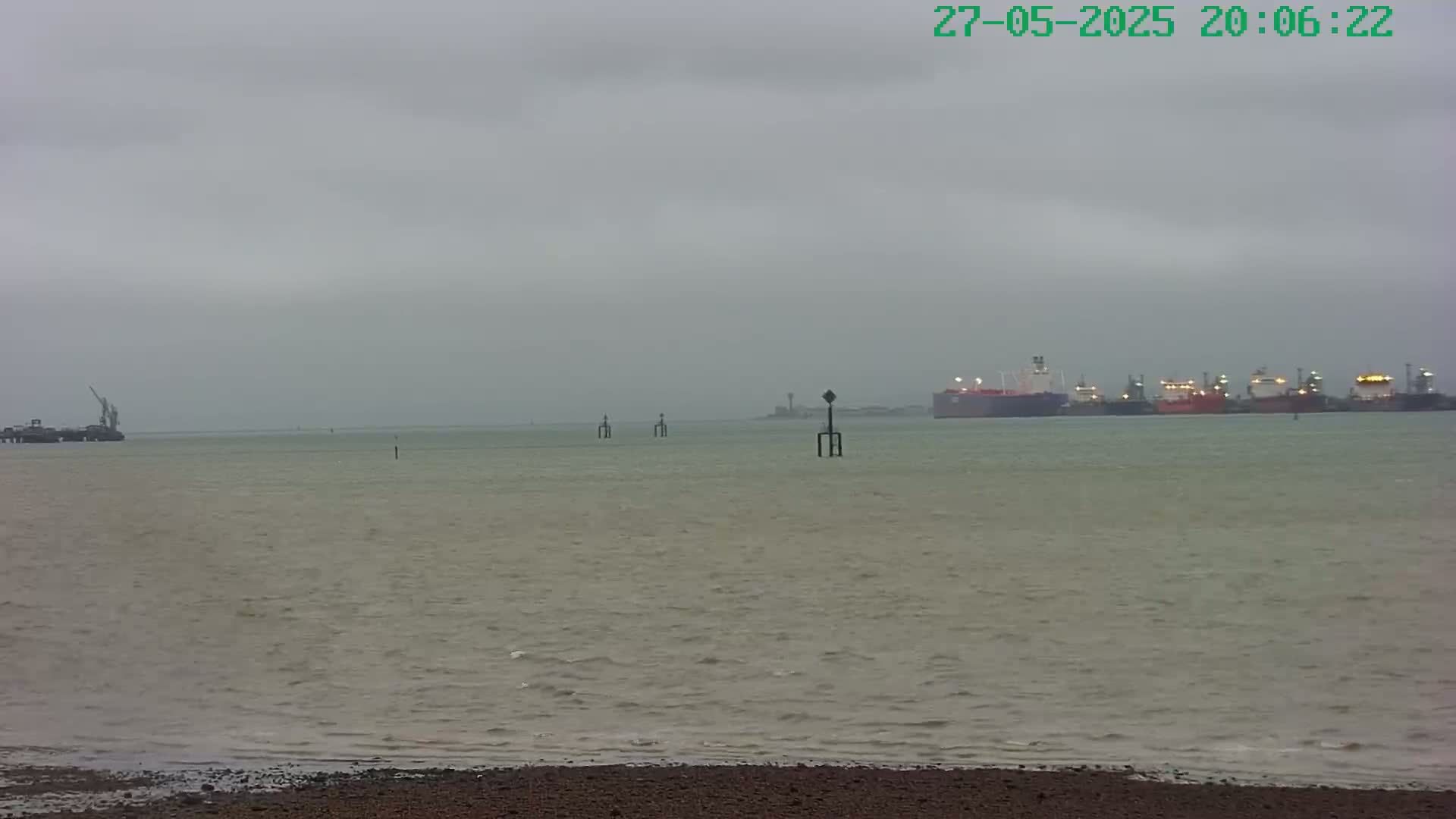 A gray, overcast day shows a body of water with several ships in the distance, near a shoreline with dark sand or pebbles.
