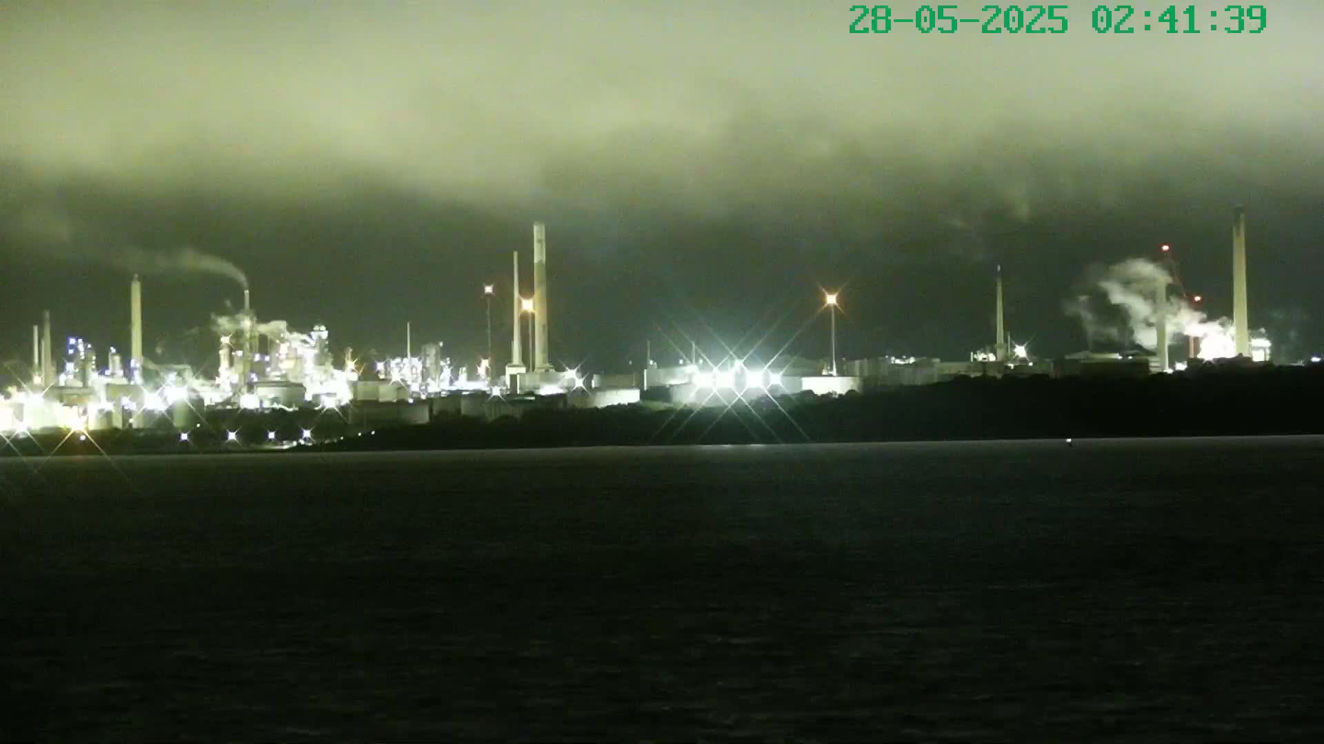 A brightly lit industrial complex is seen across a dark body of water under a cloudy night sky.