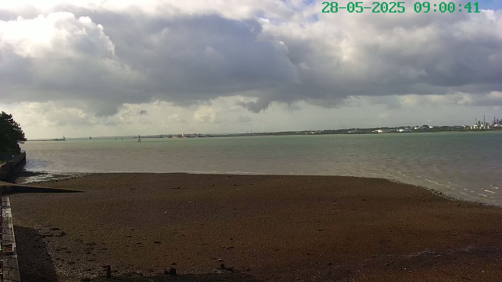 A mostly cloudy day reveals a wide expanse of dark-brown mudflats beside a calm body of water, with a distant industrial shoreline visible under a mostly cloudy sky.