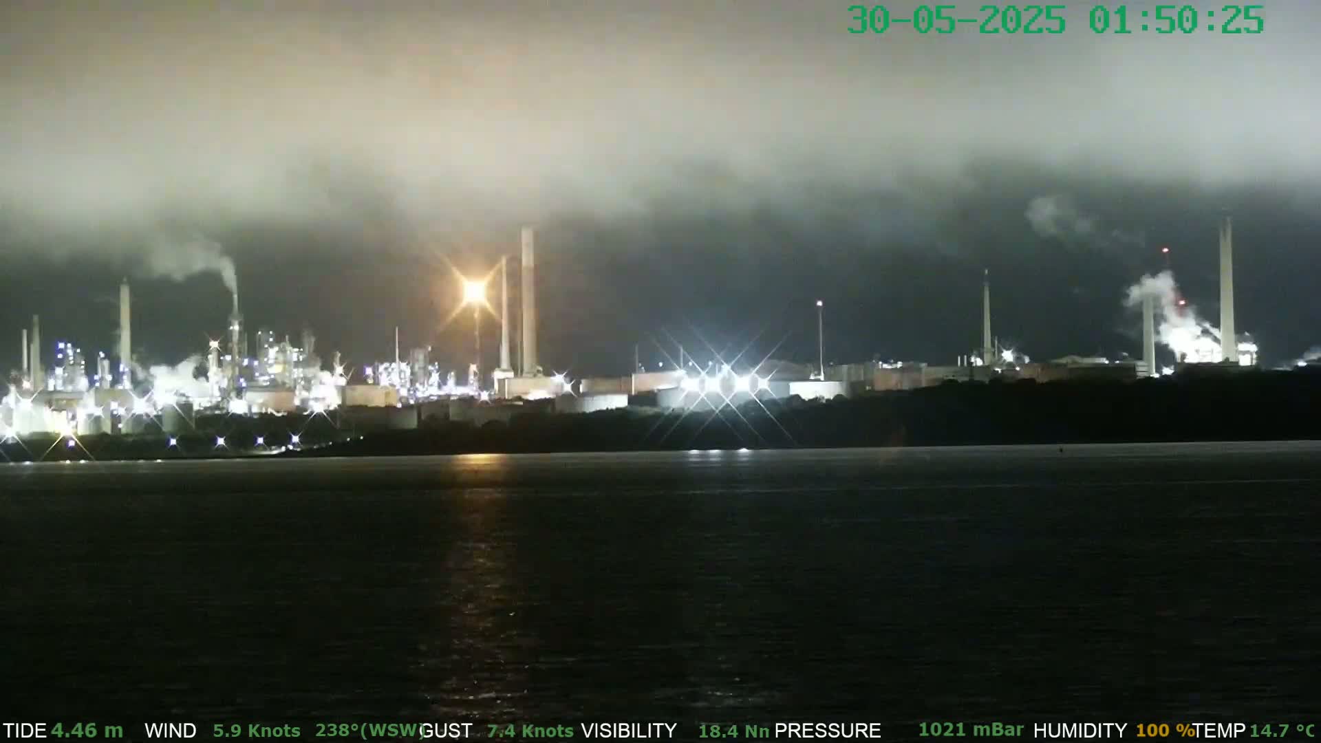 A nighttime view of a brightly lit industrial plant emitting smoke under a foggy sky, across a dark body of water.