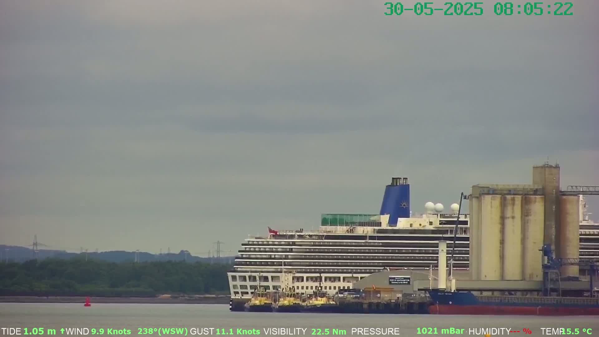 A large cruise ship is docked near industrial grain silos on an overcast day with a moderate wind.