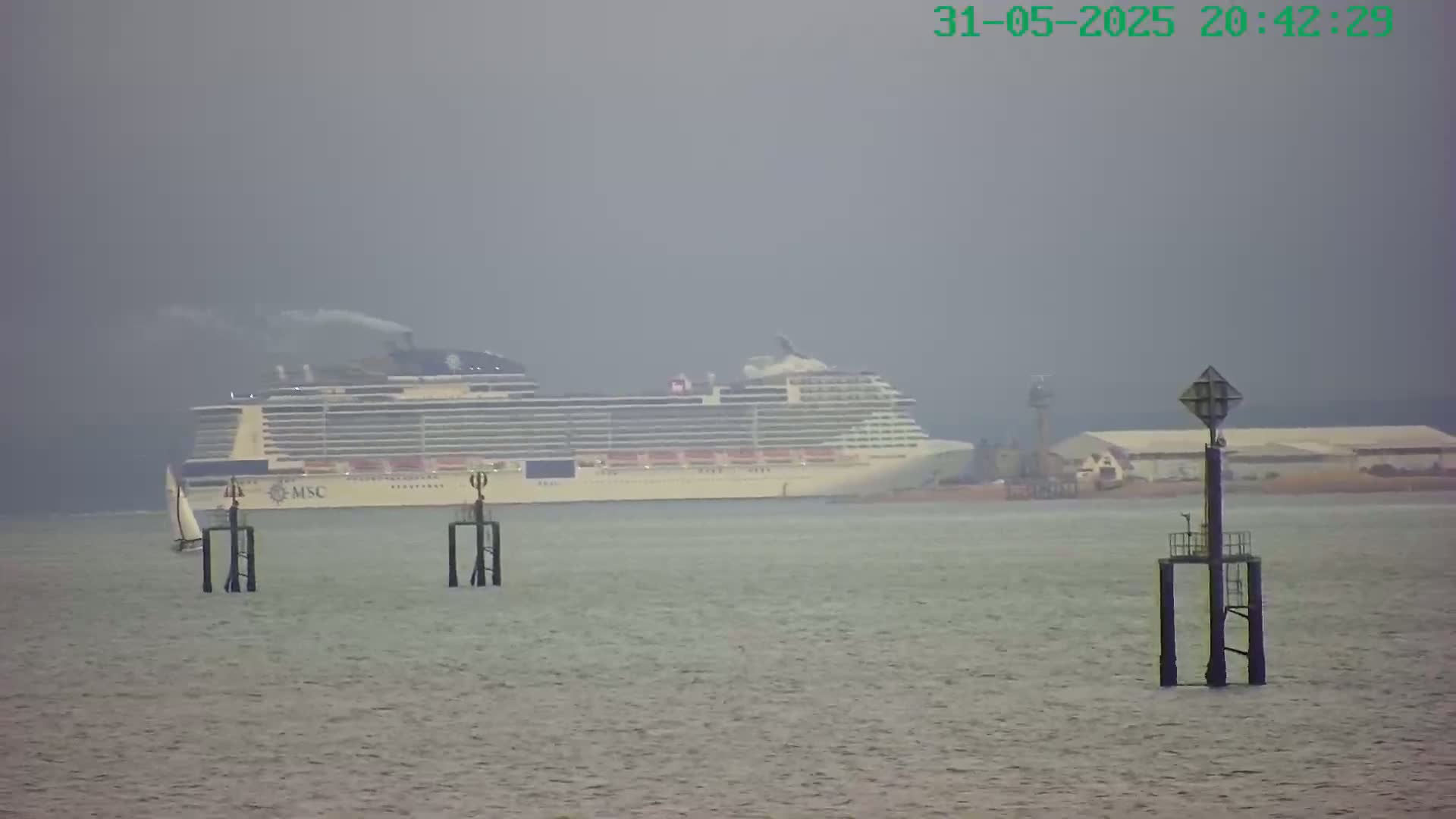 A large cruise ship is visible in the distance on a hazy, overcast day, with two navigation markers in the foreground and a small sailboat to the left.
