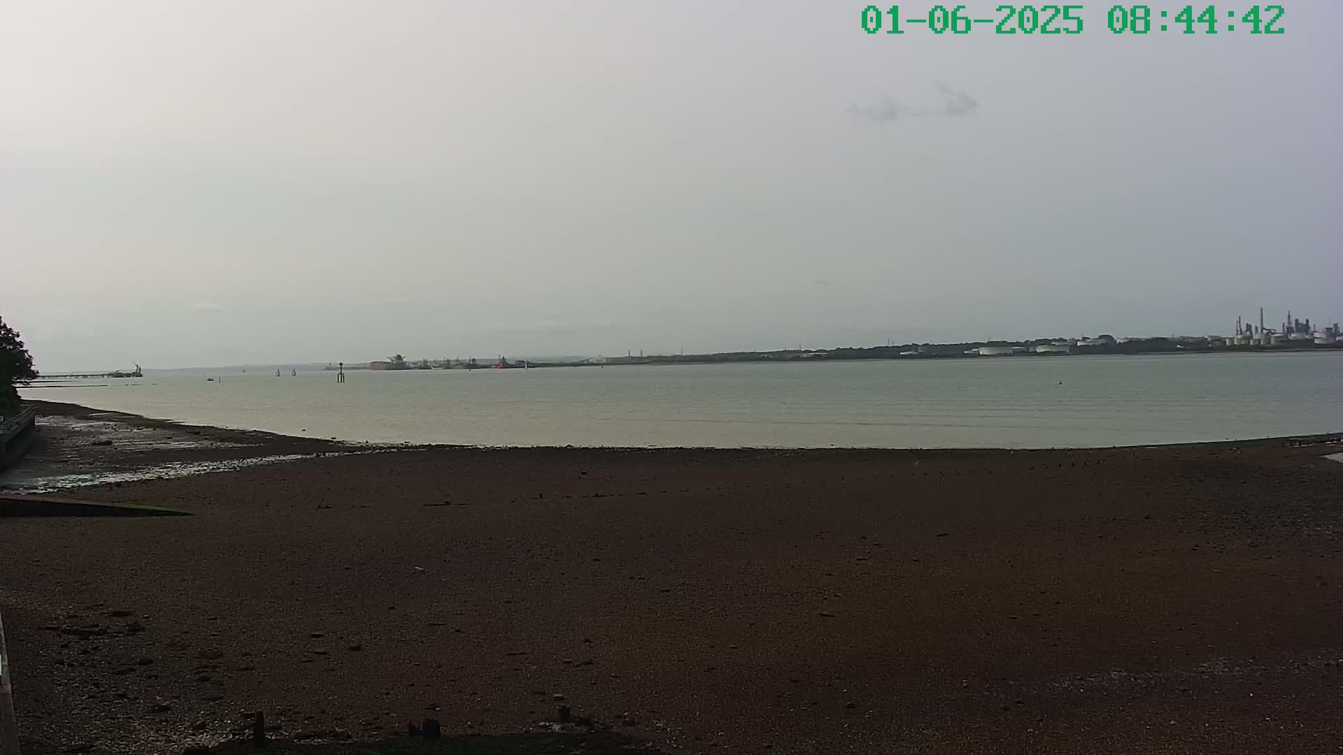 A wide shot of a dark, muddy shore receding from a calm, light-grey body of water under an overcast sky, with industrial structures visible in the distance.