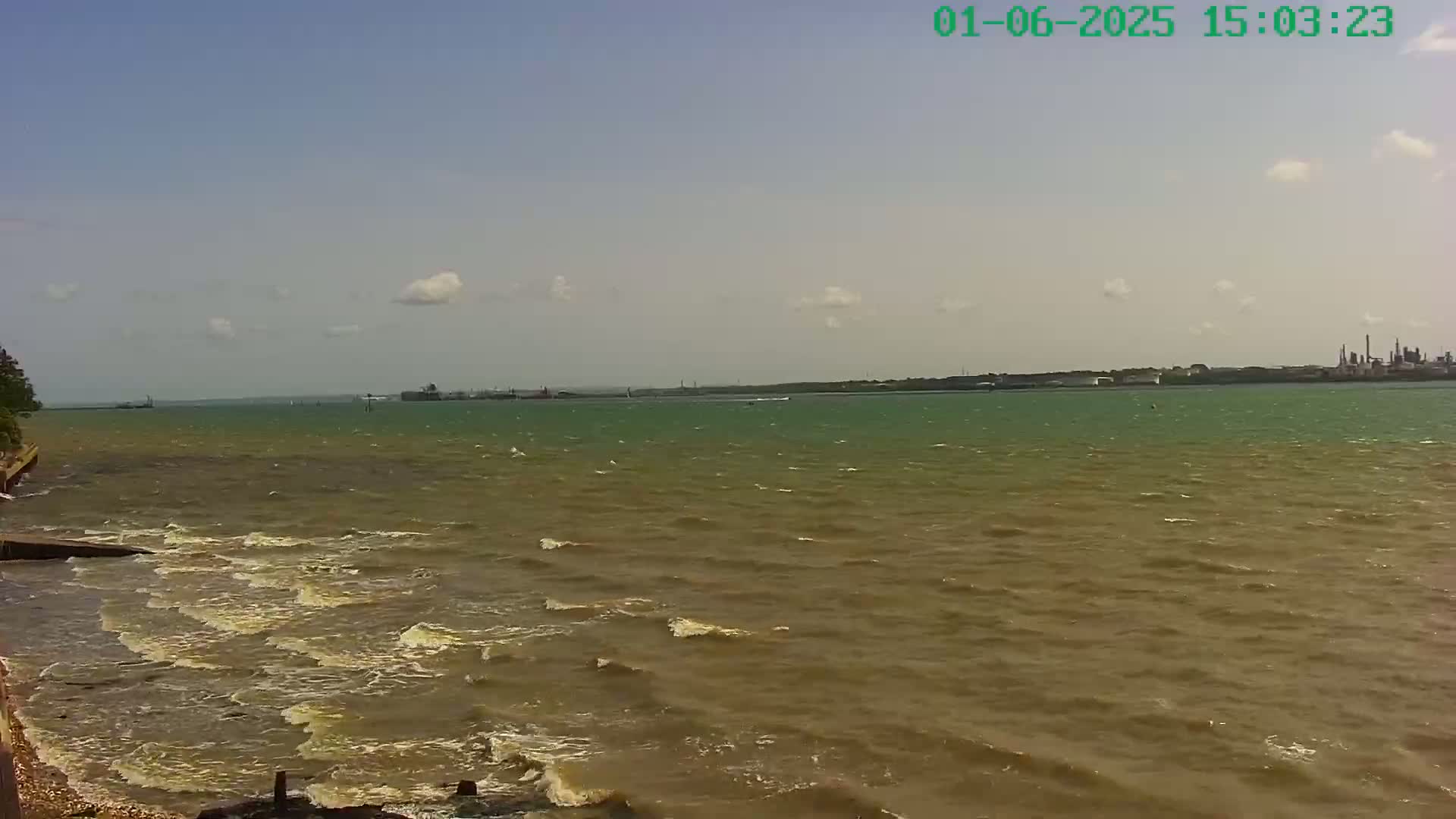 A partly cloudy sky overlooks a choppy, brownish-green body of water with a distant industrial shoreline.