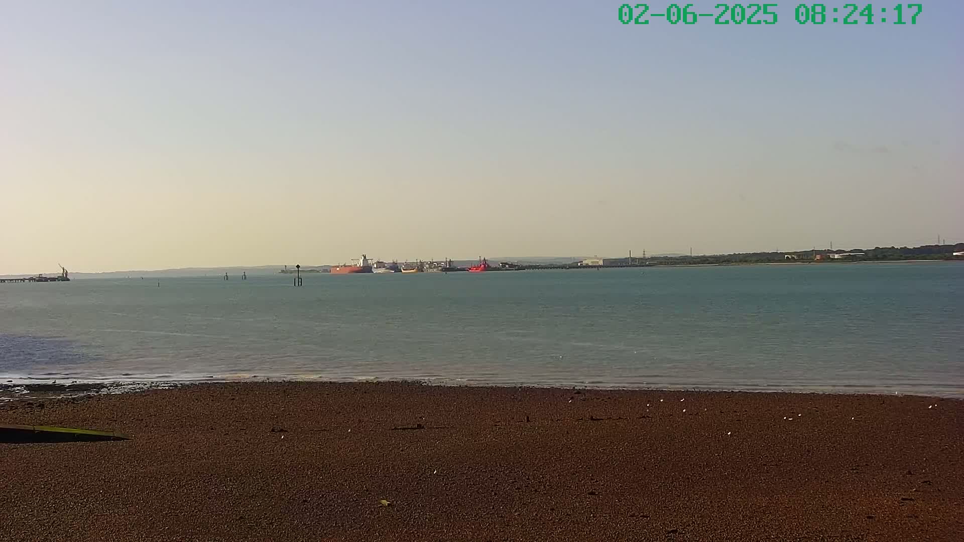 A calm, light-grey body of water stretches out to a distant shoreline under a clear, pale sky, with several ships visible in the distance and a dark brown, pebbly beach in the foreground.