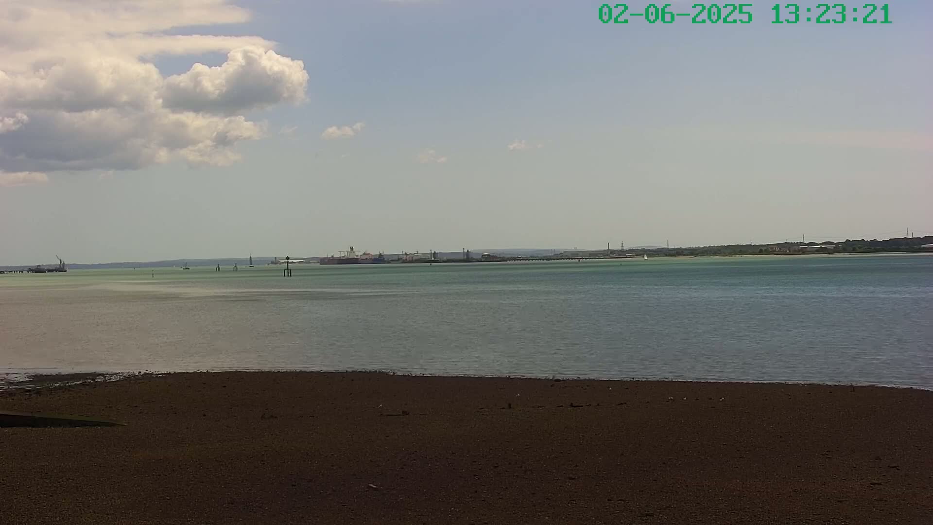 A partly cloudy day reveals a view of a calm body of water with a dark-colored sandy shore in the foreground, and a distant industrial waterfront.