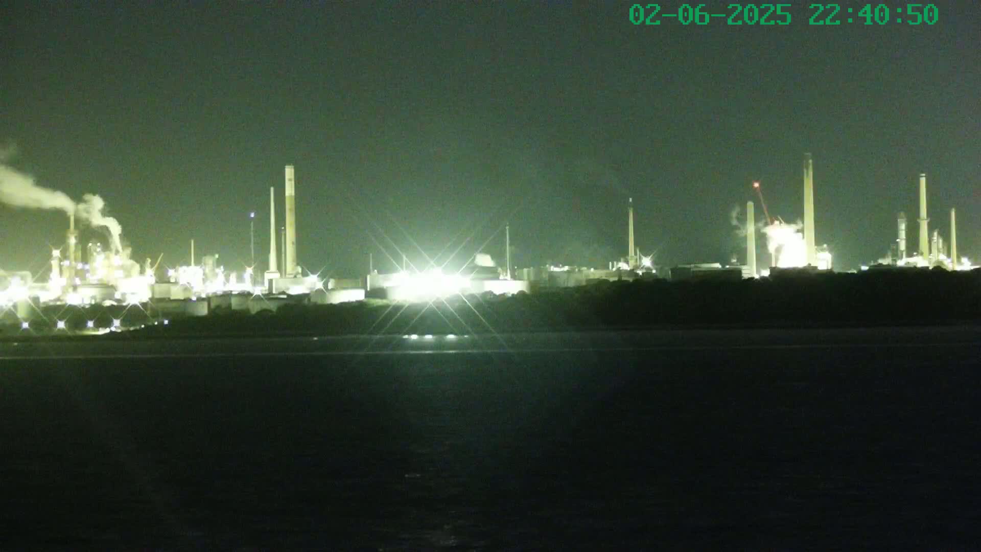 A brightly lit industrial complex at night is seen across a dark body of water under a dark sky.