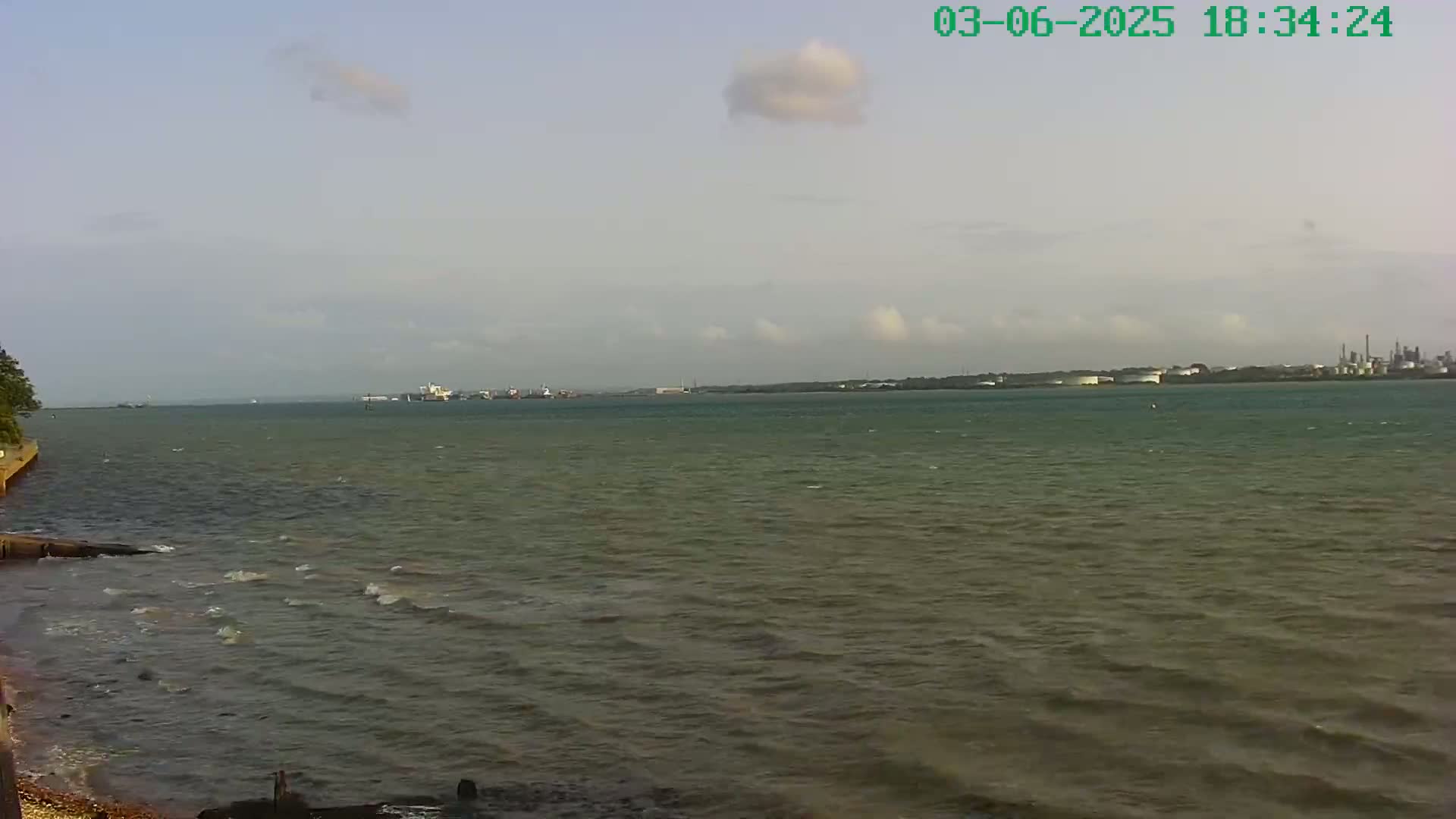 A partly cloudy day reveals a wide body of water with small waves lapping a shoreline, and an industrial area visible in the distance.