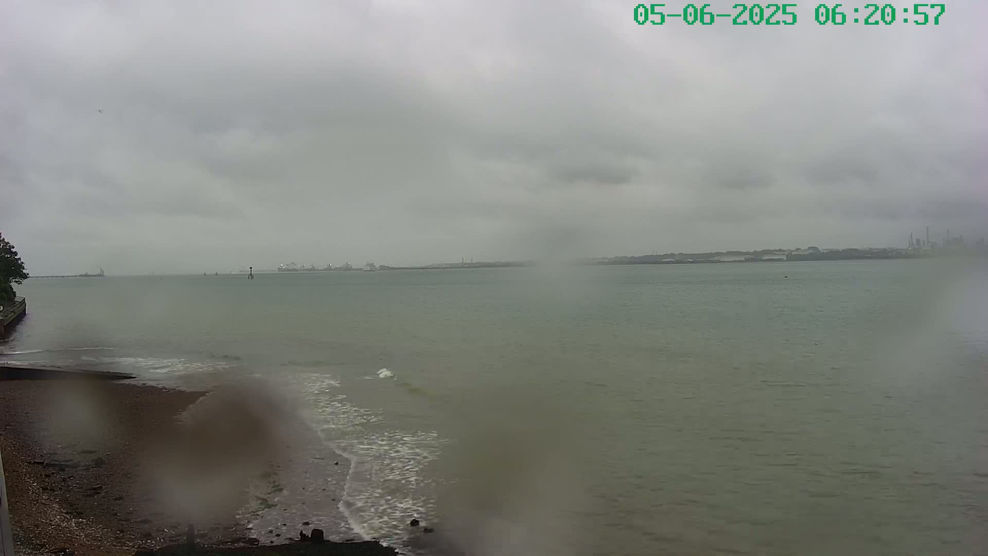 A grey, overcast day shows a view of a calm body of water with a shoreline and distant industrial structures.