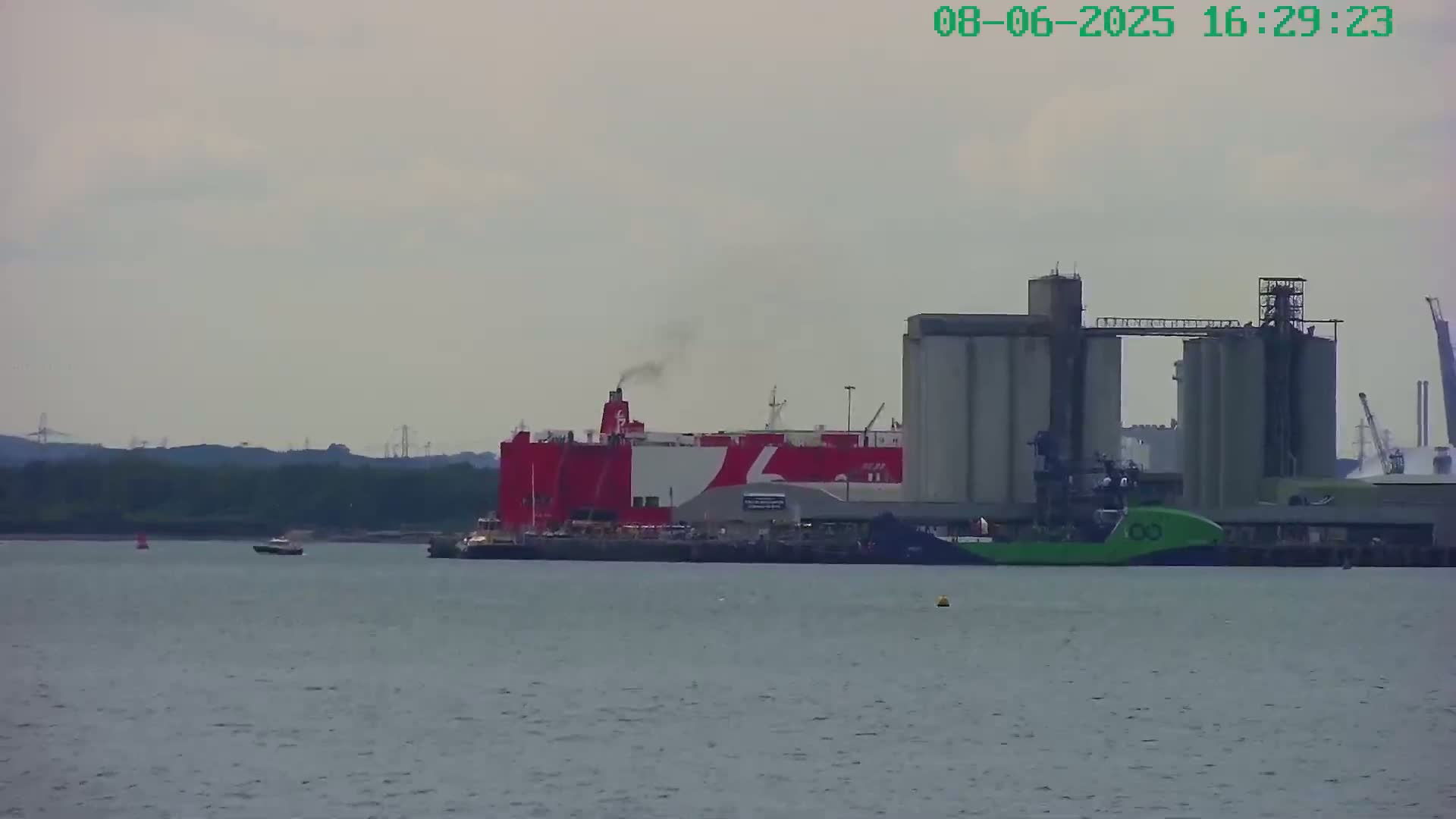 A large red and white ship is docked near a cluster of industrial silos on a calm, overcast day.