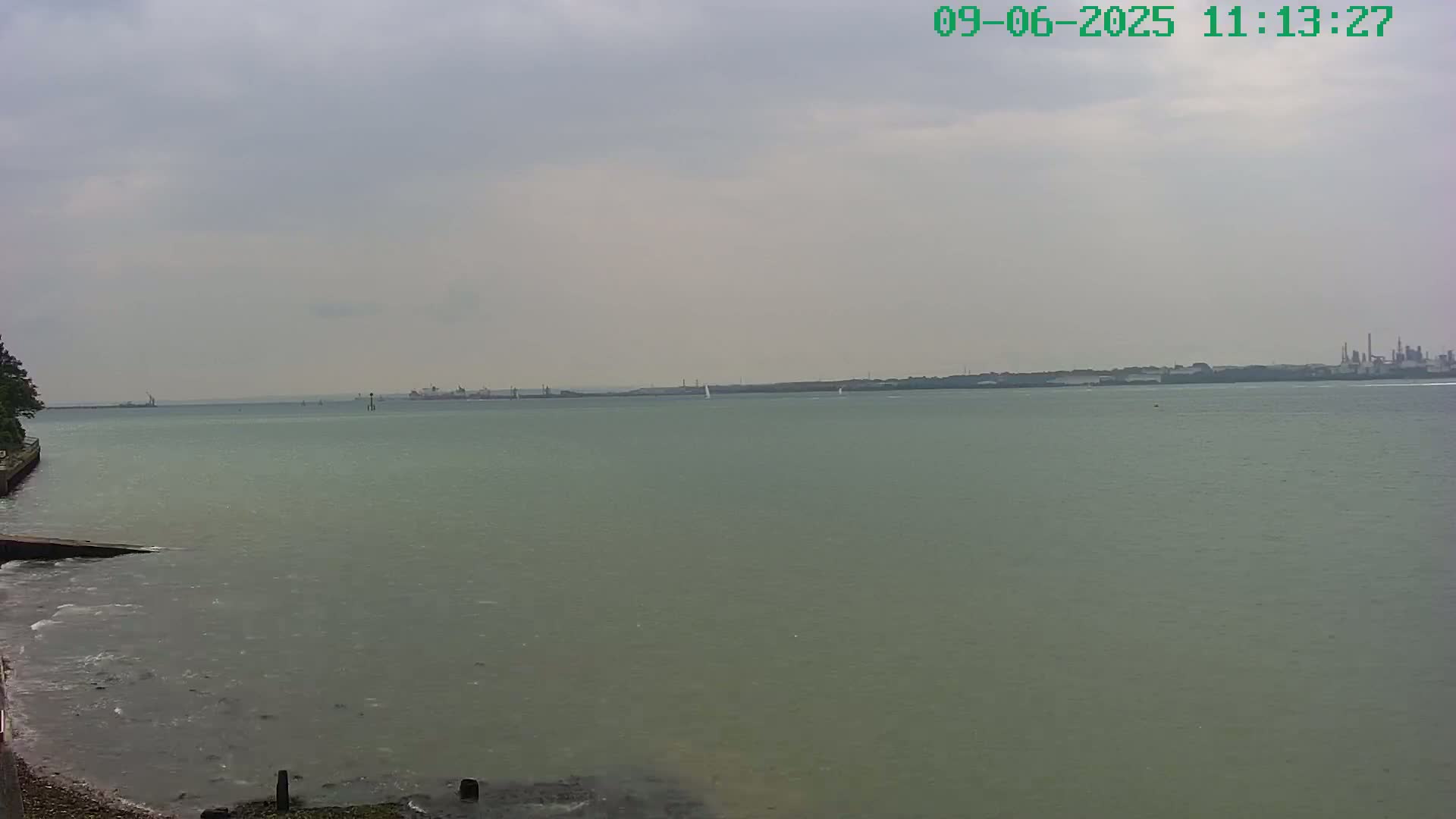 A calm, overcast day shows a wide expanse of grey-green water with a distant shoreline featuring industrial structures.