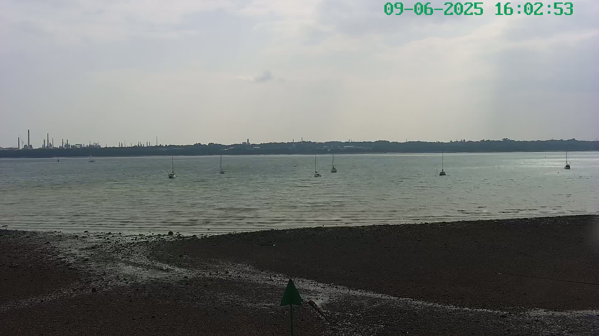 A dark, sandy shore leads to a calm, gray body of water with several sailboats, and a distant industrial skyline under an overcast sky.