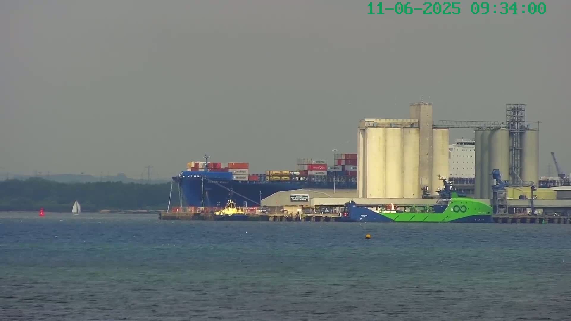 A large container ship is docked at an industrial port with grain silos under hazy skies, and a small sailboat is visible in the distance.