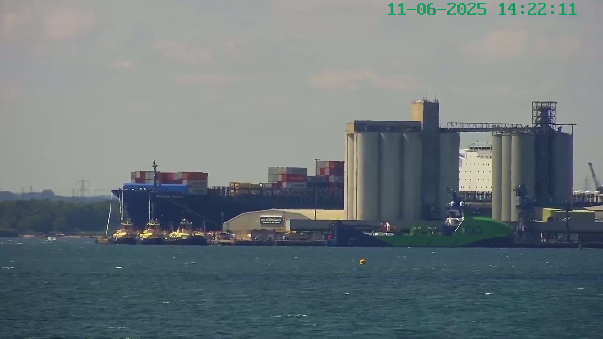 A large container ship is moored at a dock next to several grain silos and tugboats, under a hazy, sunny sky.