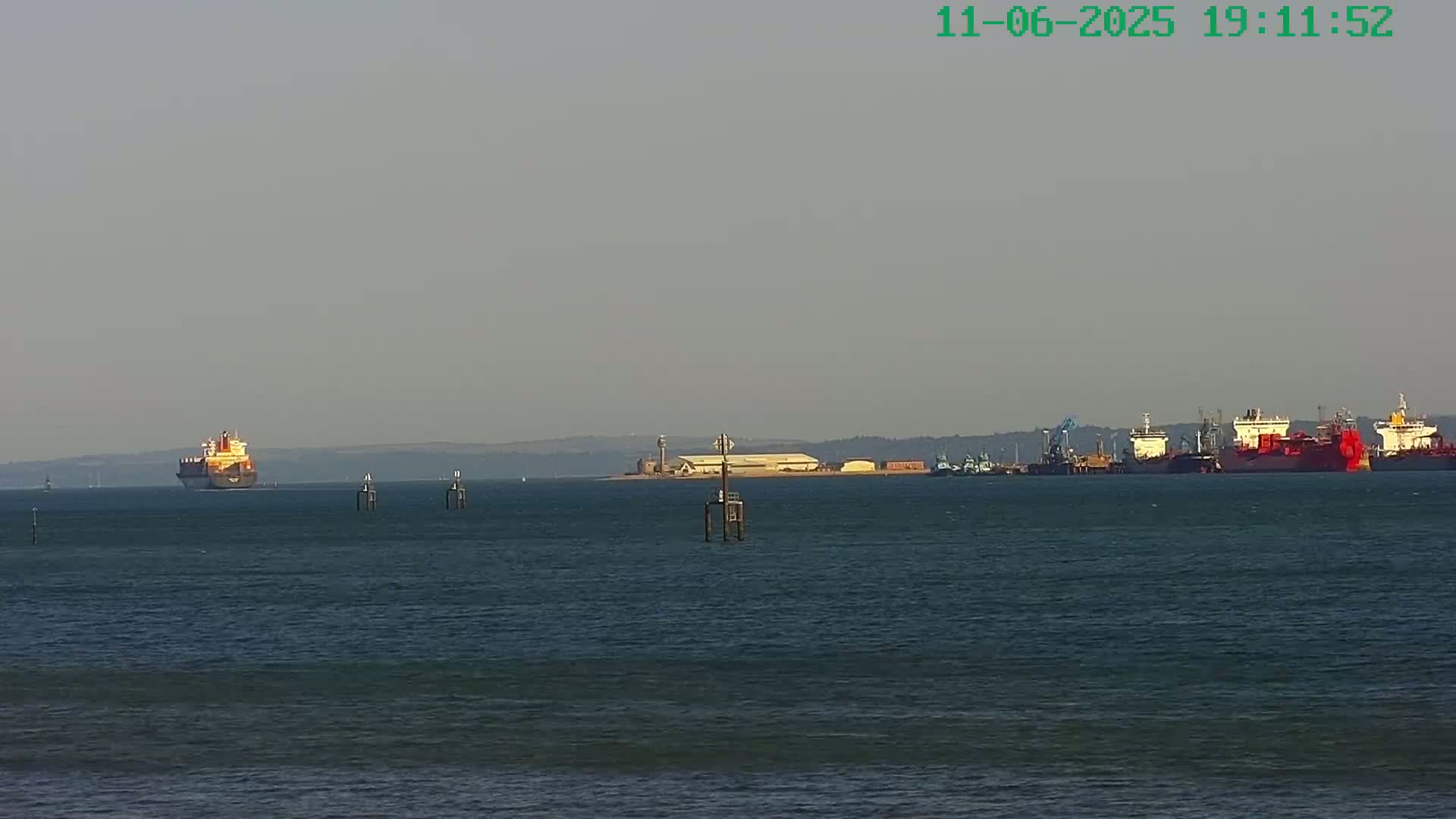 A large cargo ship is seen sailing on calm water toward a port where several other ships are docked under a hazy sky.