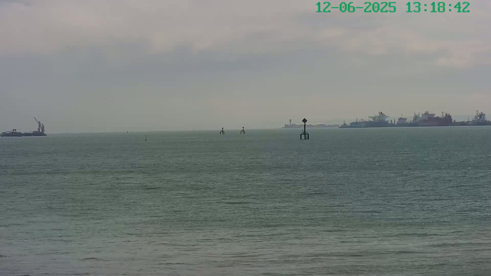 A calm, gray day shows a wide expanse of gray-green water with several ships and navigational markers in the distance.