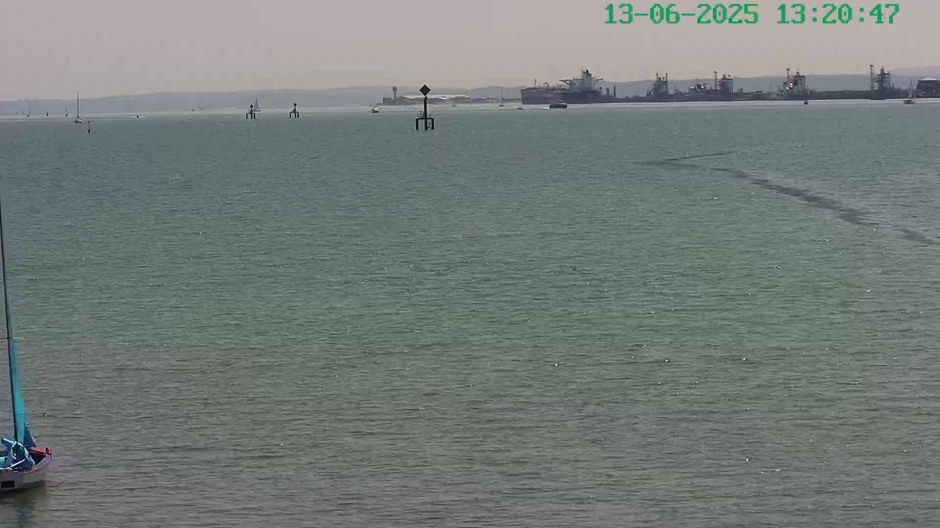 A sailboat is in the foreground of a calm, gray-green body of water on a hazy day, with several other boats and a large industrial area visible in the distance.