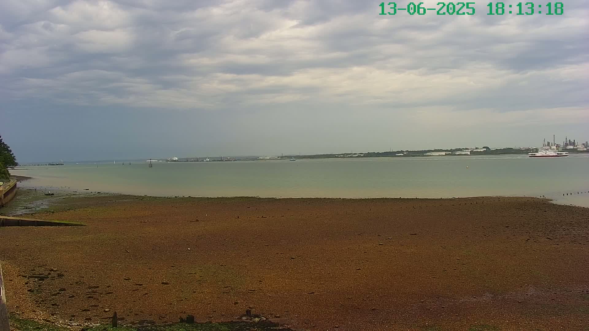 A wide shot of a mostly exposed, brown, pebbly shoreline on a cloudy day, with calm, light-green water extending to a distant industrial area.