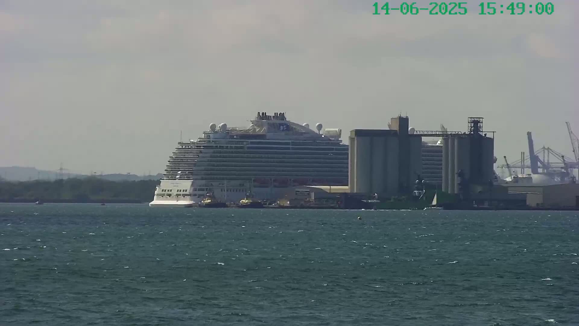 A large cruise ship is docked near an industrial area on a partly cloudy day.