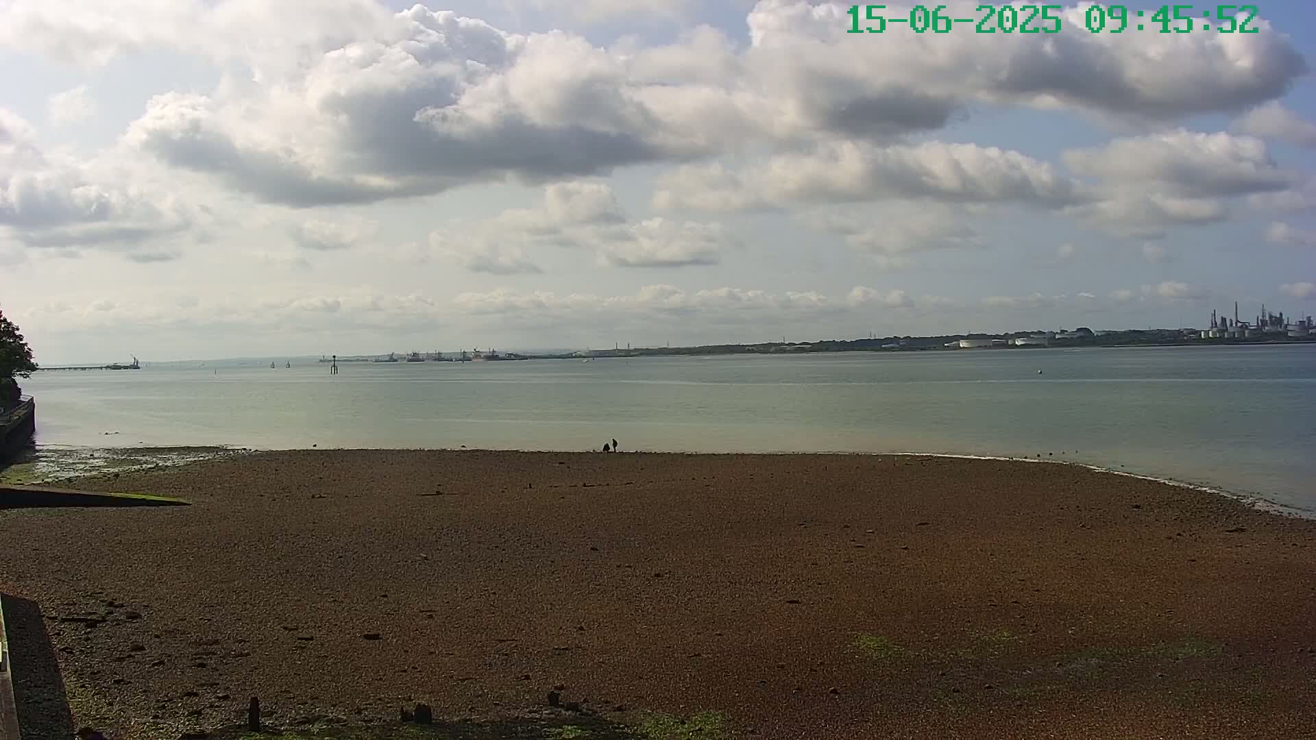 A partly cloudy day reveals a wide expanse of calm water, a flat, brown, pebbly shore in the foreground, and distant industrial structures on the horizon.