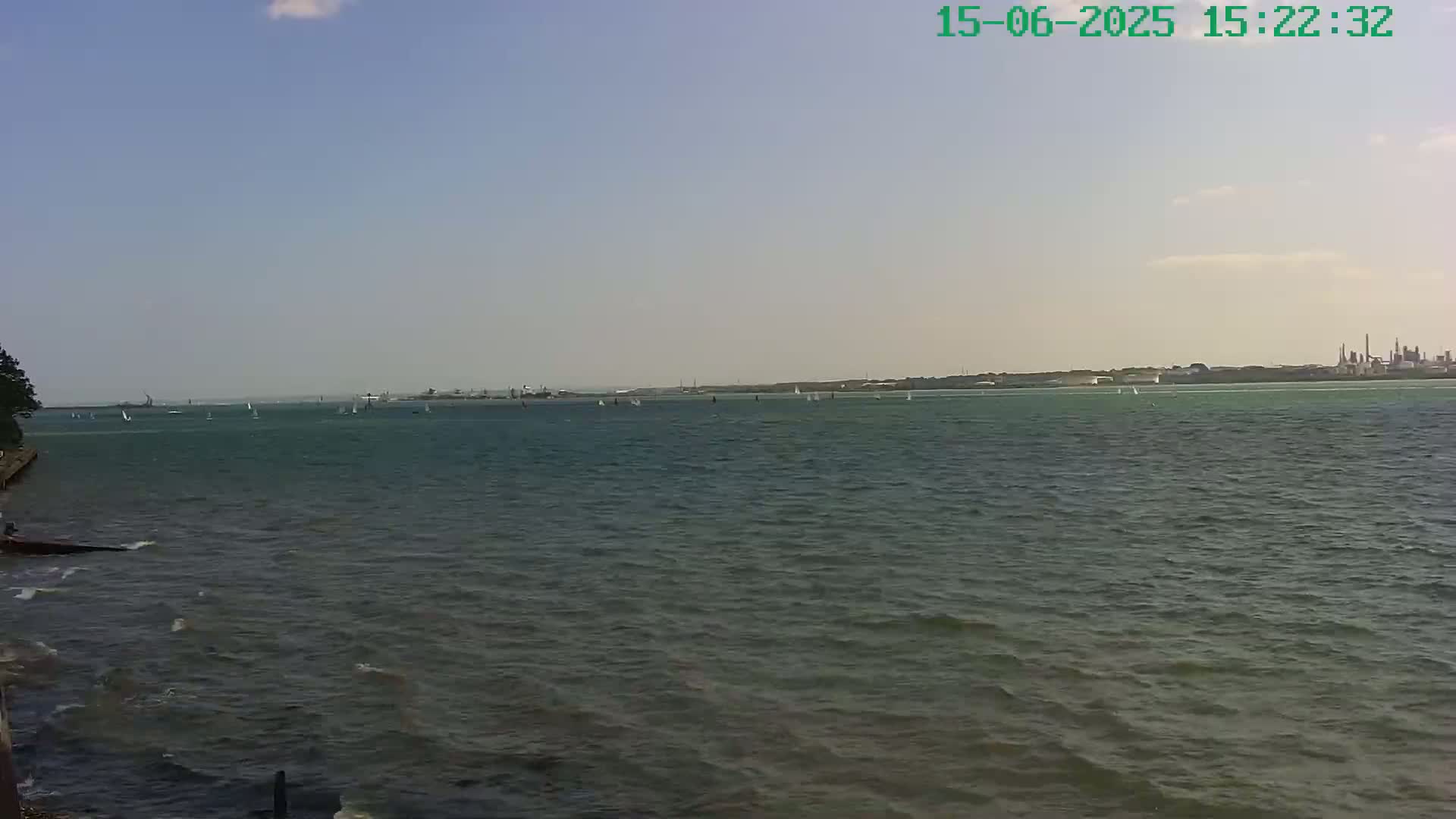 A partly cloudy day shows a body of water with many sailboats and a distant industrial shoreline.