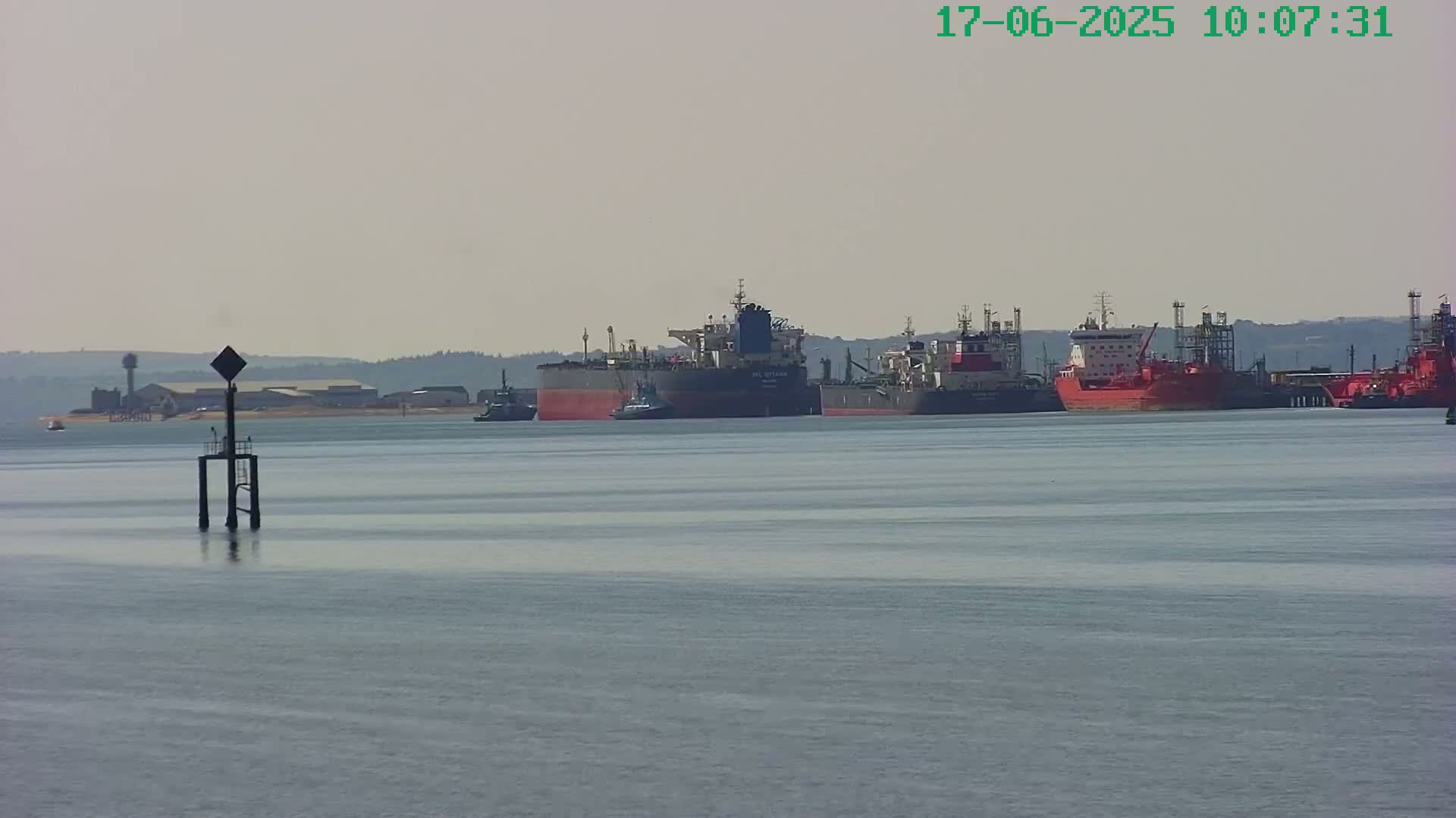 Several large ships are at anchor in calm water, with a hazy sky and distant shoreline visible.