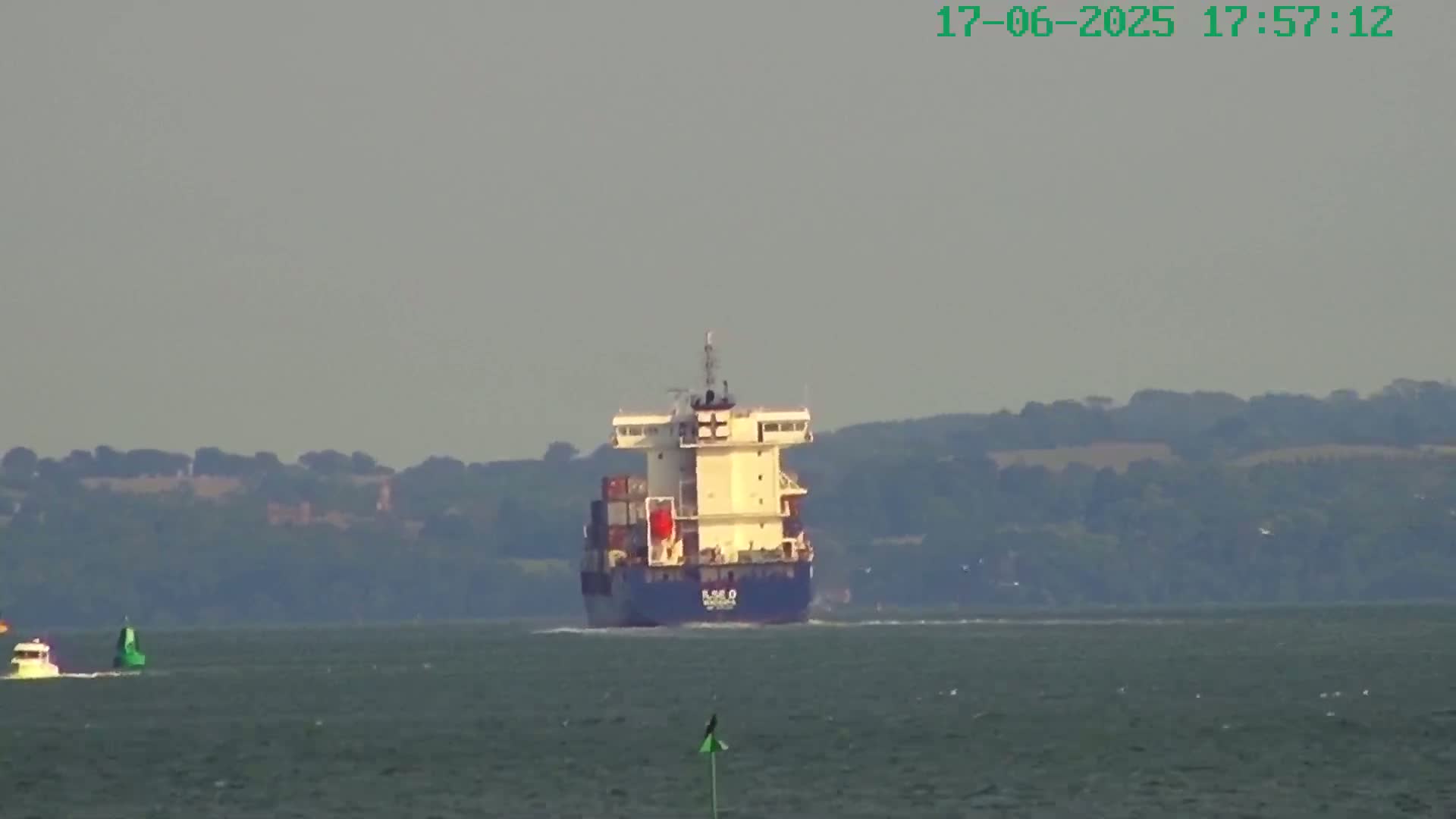 A large container ship sails on a calm, hazy sea toward a distant, tree-covered shoreline.