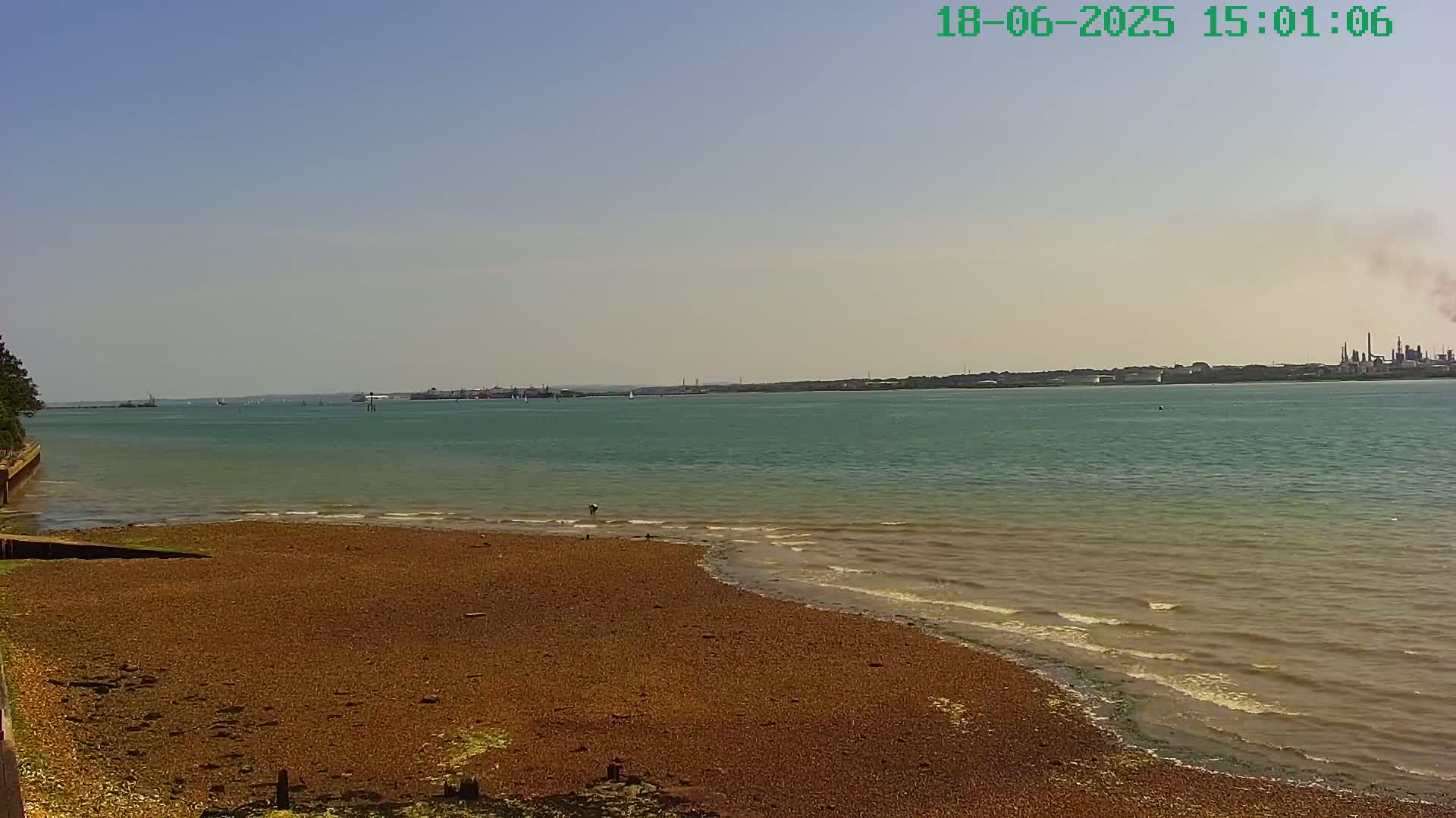 A calm, sunny day reveals a pebble beach meeting a wide body of water with distant industrial structures and a few small vessels visible on the horizon.