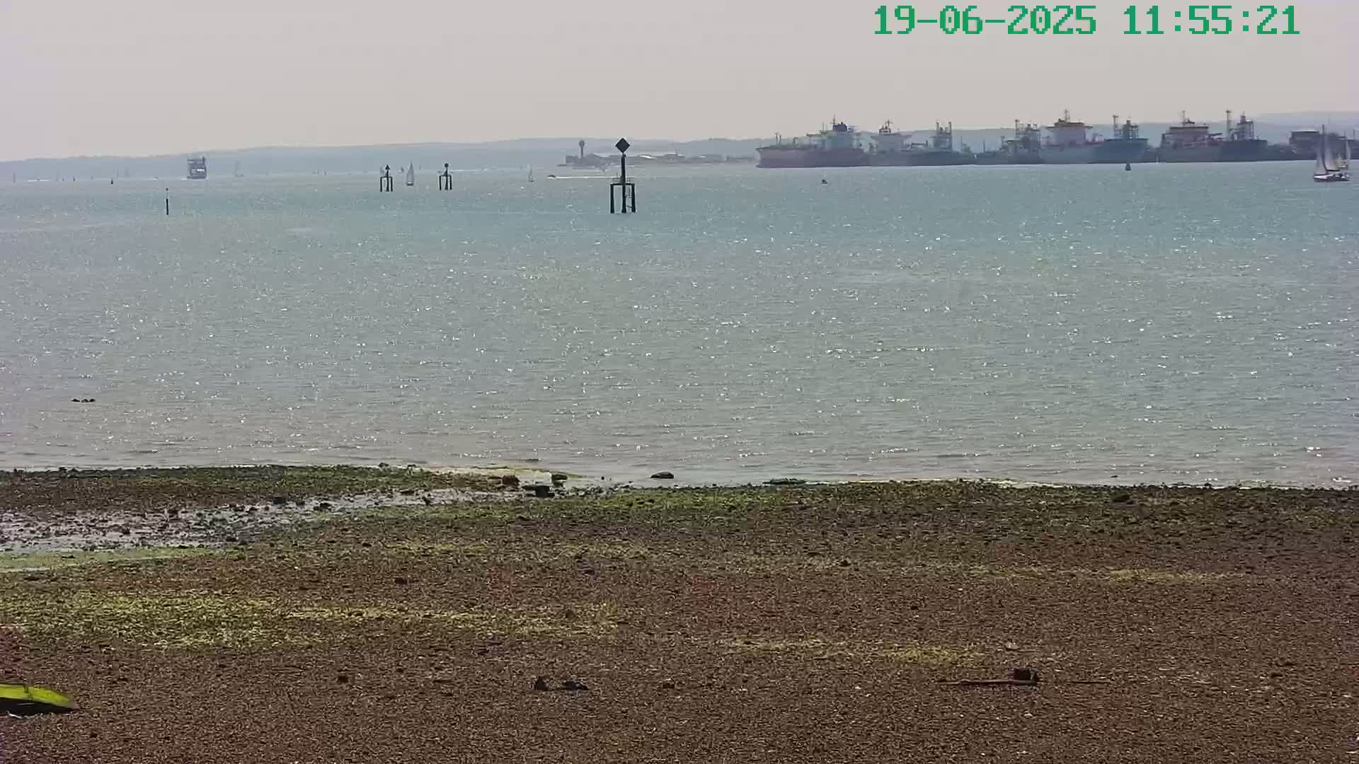 A rocky shoreline stretches across the bottom of the image, meeting a calm, sunlit body of water where several ships are visible in the distance under a hazy sky.