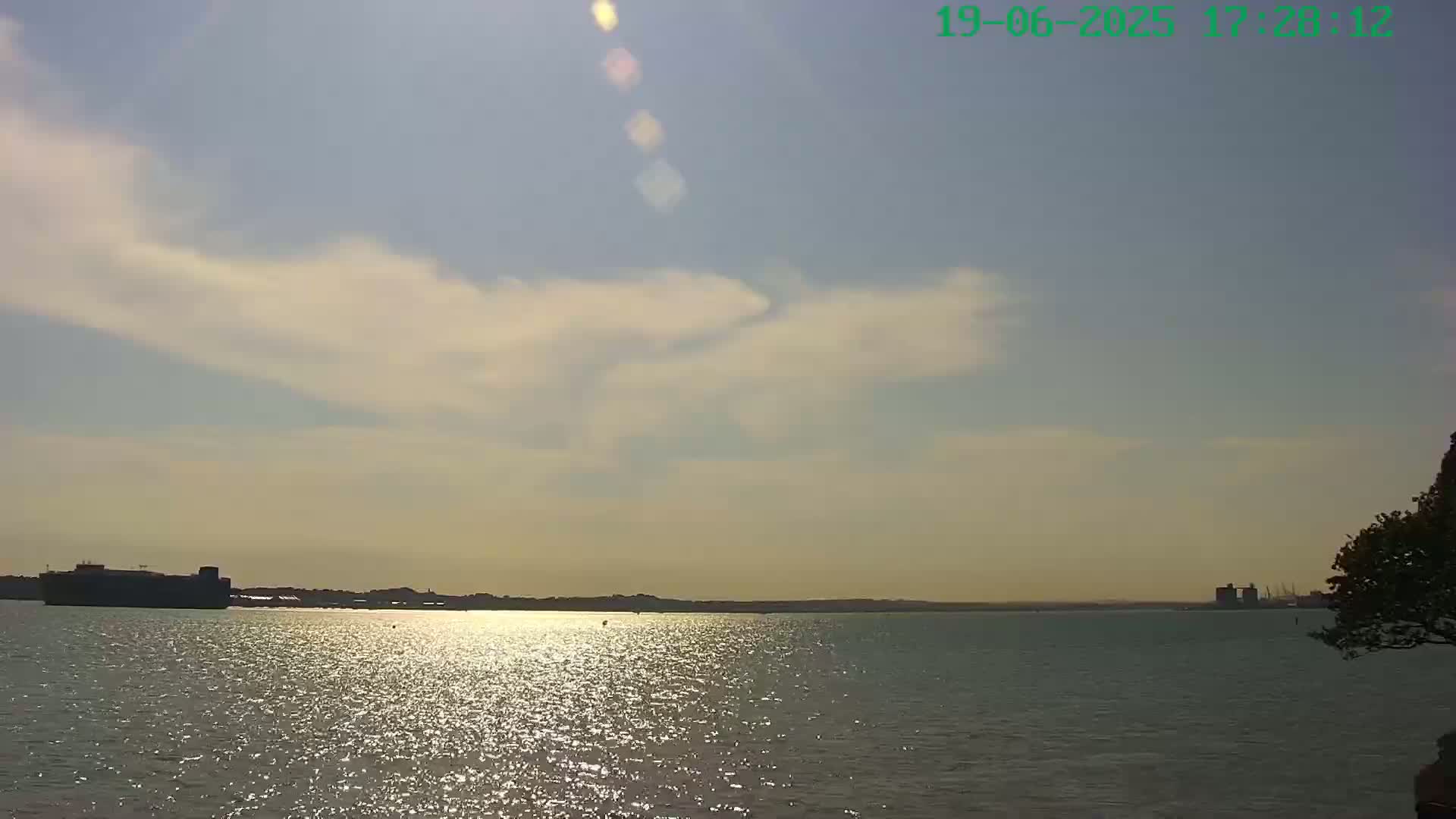 A partly cloudy day shows a calm body of water reflecting sunlight, with a distant shoreline featuring some structures and a tree partially visible in the lower right corner.