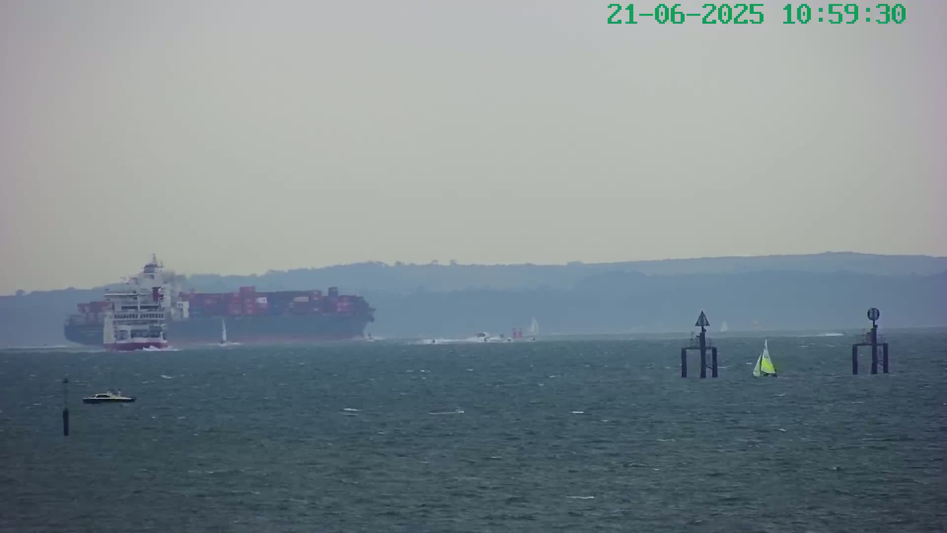 A large container ship and several smaller vessels are visible on a hazy, gray day at sea, with a distant shoreline in the background.