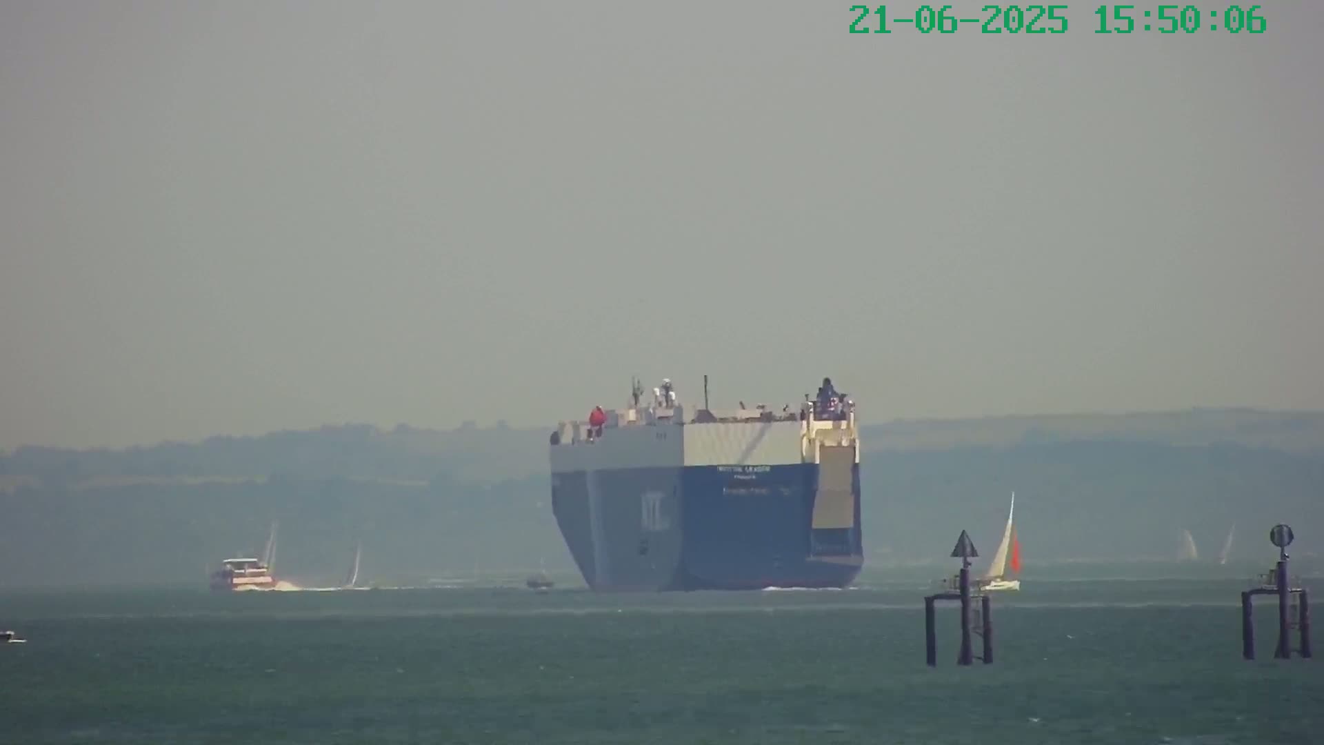 A large cargo ship is on the water under hazy conditions, with several smaller boats and sailboats in the distance.