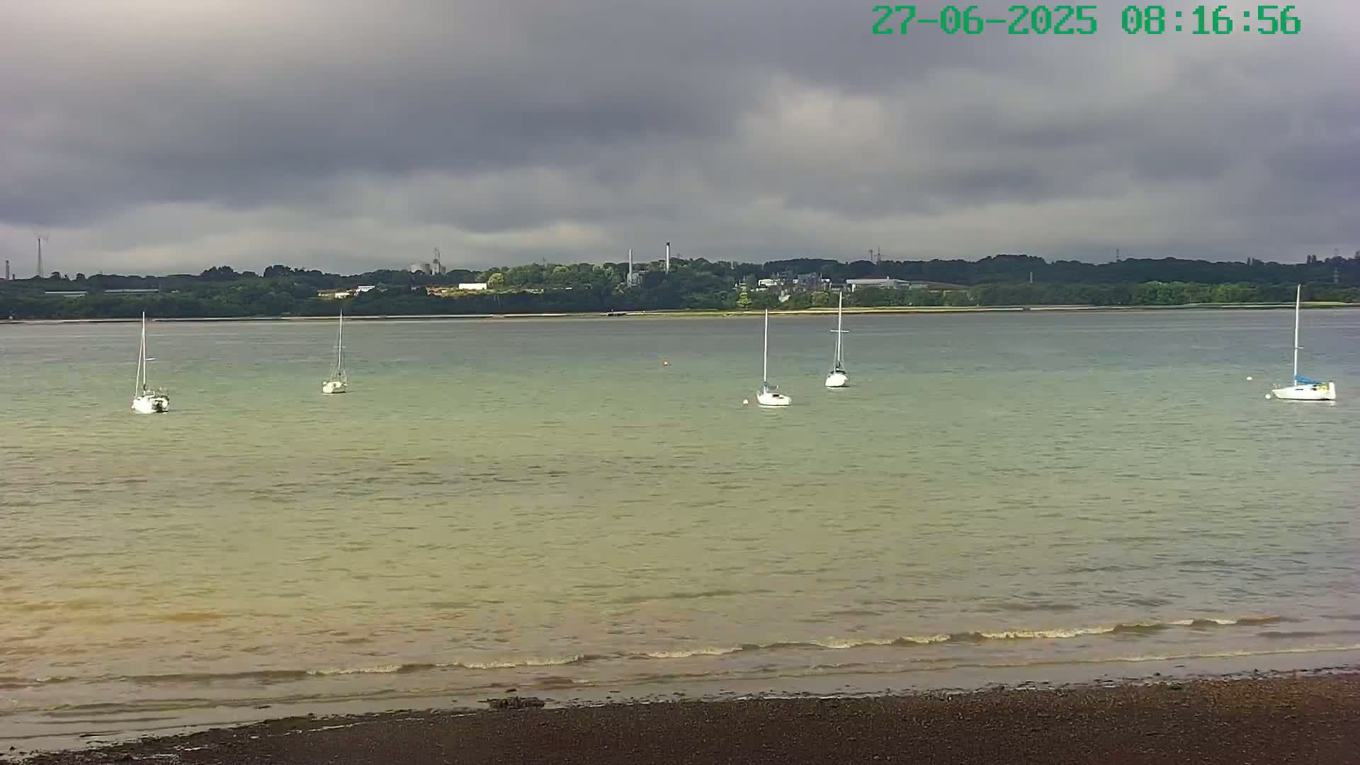 Several sailboats are anchored in calm, light-green water under a cloudy sky, with a distant shoreline visible.