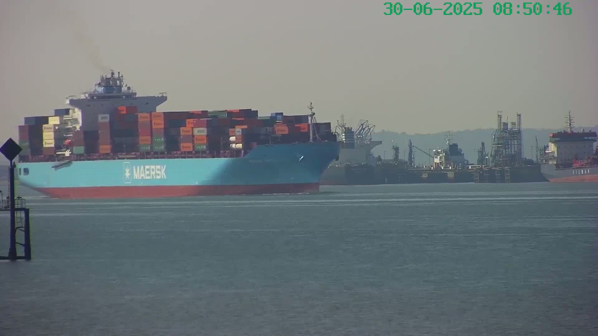 A large container ship, labeled Maersk, sails on calm water past several other ships and industrial structures on a hazy day.