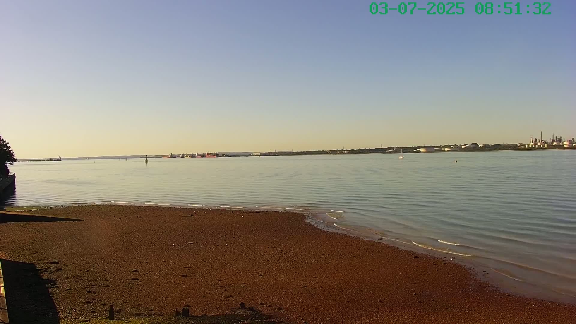 A calm, clear day reveals a sandy shore, gentle waves lapping at the beach, and a distant industrial area across a body of water.