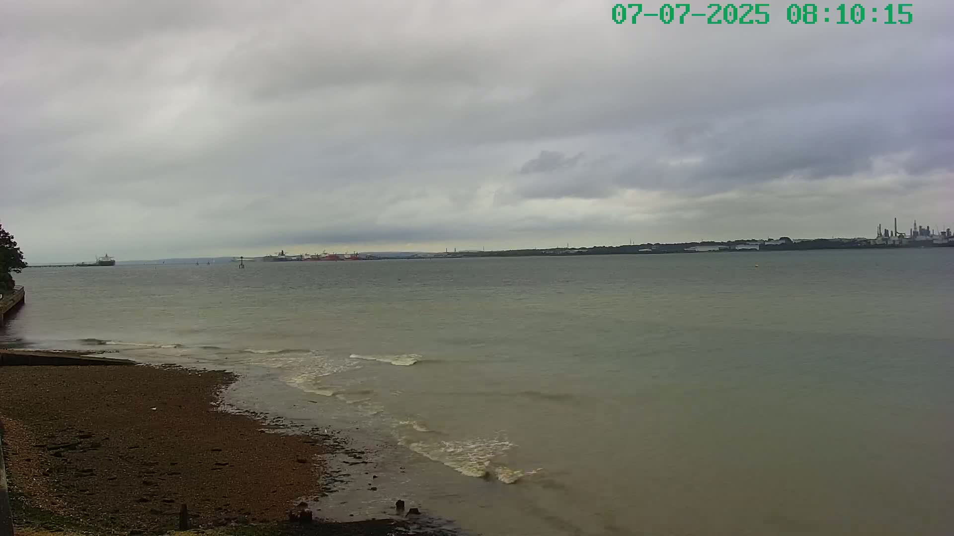 A pebbly shore meets a calm, grey body of water under an overcast sky, with distant ships and industrial structures visible on the horizon.