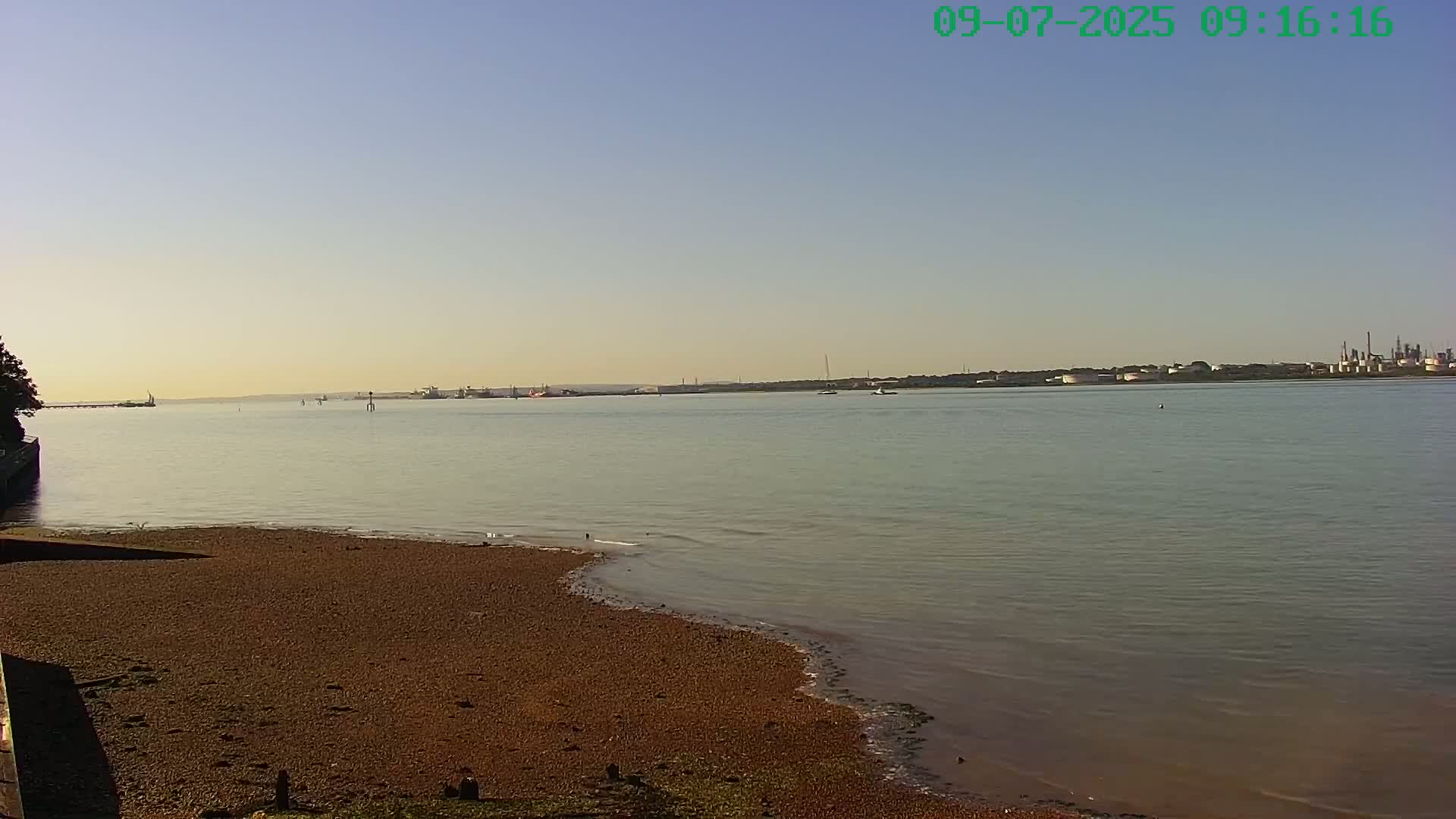 A calm, light-colored body of water stretches to a distant industrial area under a clear, pale sky, with a small sandy beach in the foreground.