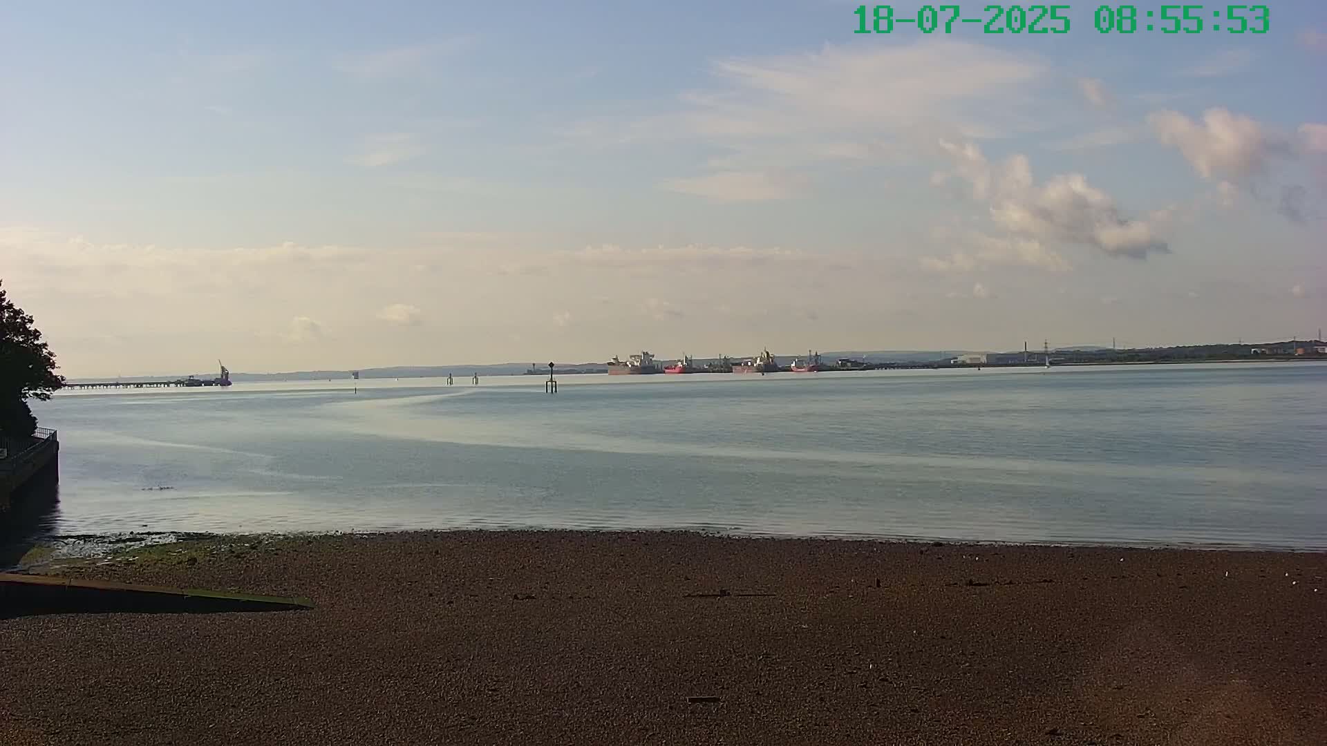 A mostly sunny day reveals a calm body of water with several ships in the distance and a sandy shoreline in the foreground.