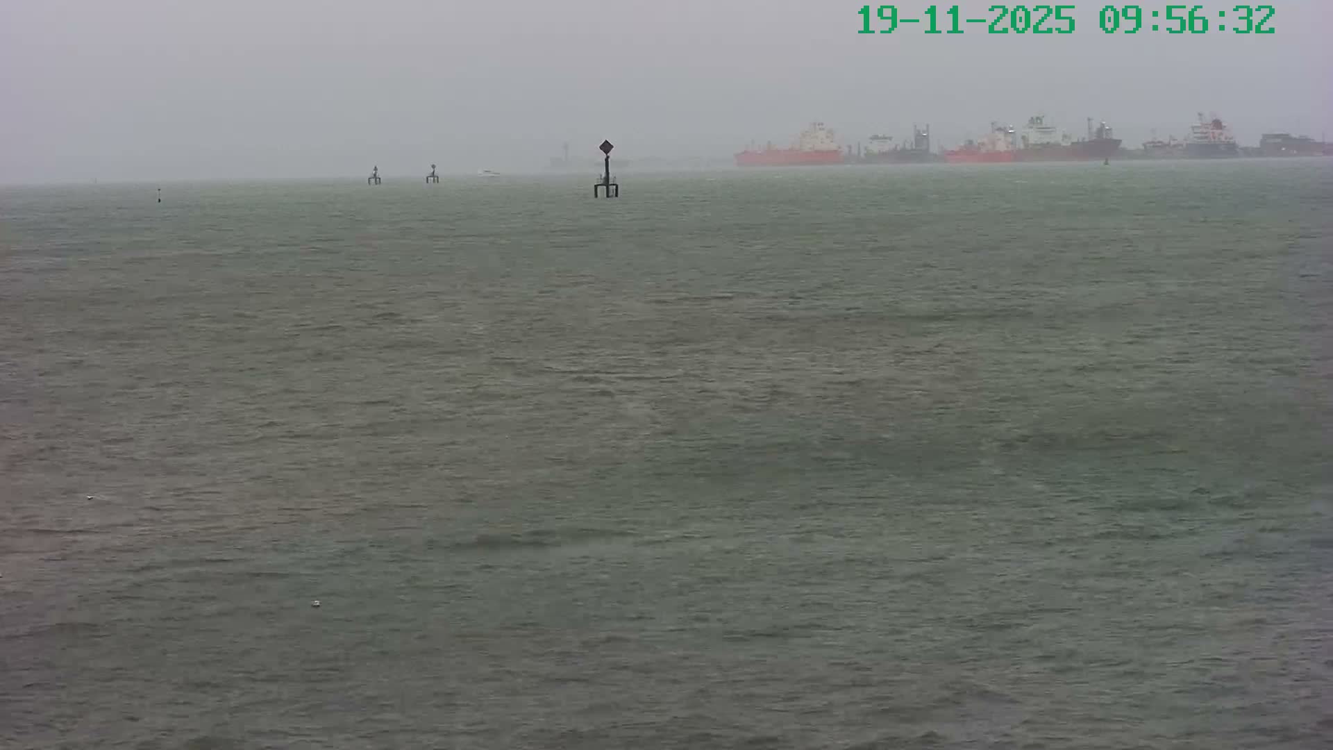 Choppy, grey-green water with several navigation markers and a small moving boat extends to a hazy background where numerous large ships are dimly visible under misty, overcast skies.