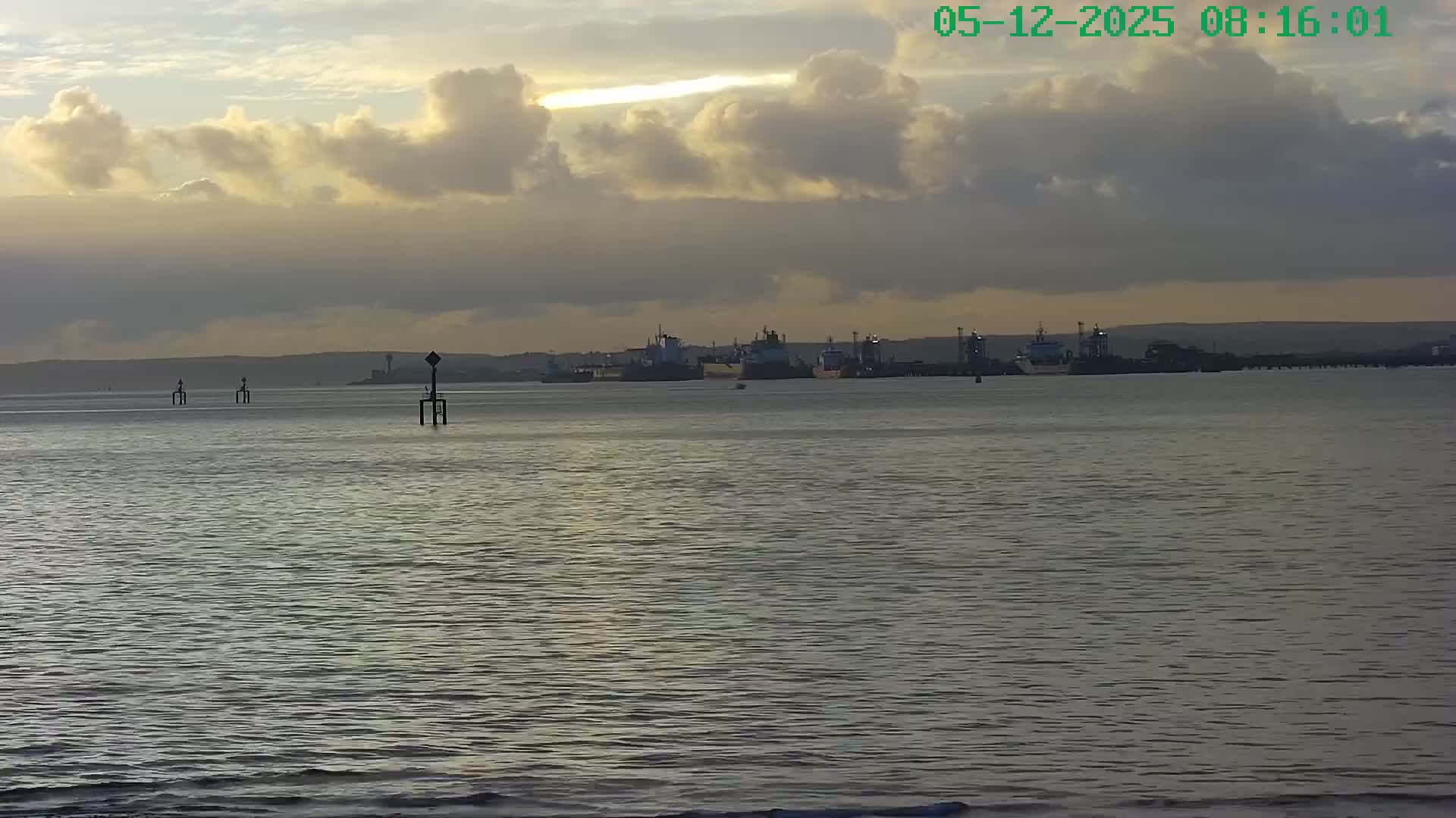 A wide view captures a calm body of water reflecting the golden light of the sun breaking through a partly cloudy sky, with several cargo ships and industrial structures visible along the distant shoreline.
