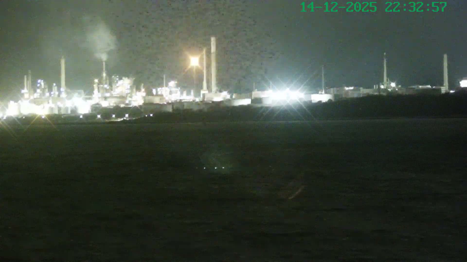 A wide view captures a calm body of water reflecting the golden light of the sun breaking through a partly cloudy sky, with several cargo ships and industrial structures visible along the distant shoreline.