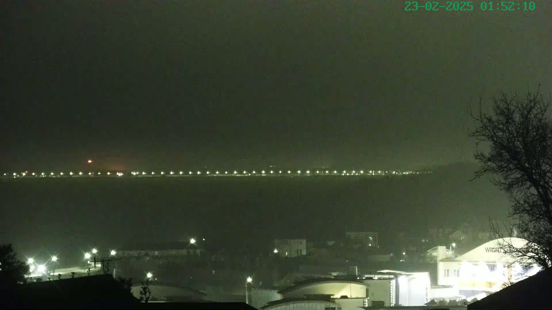 A nighttime long shot shows a line of lights across a hazy body of water, with a town and buildings in the foreground under a dark, possibly foggy sky.