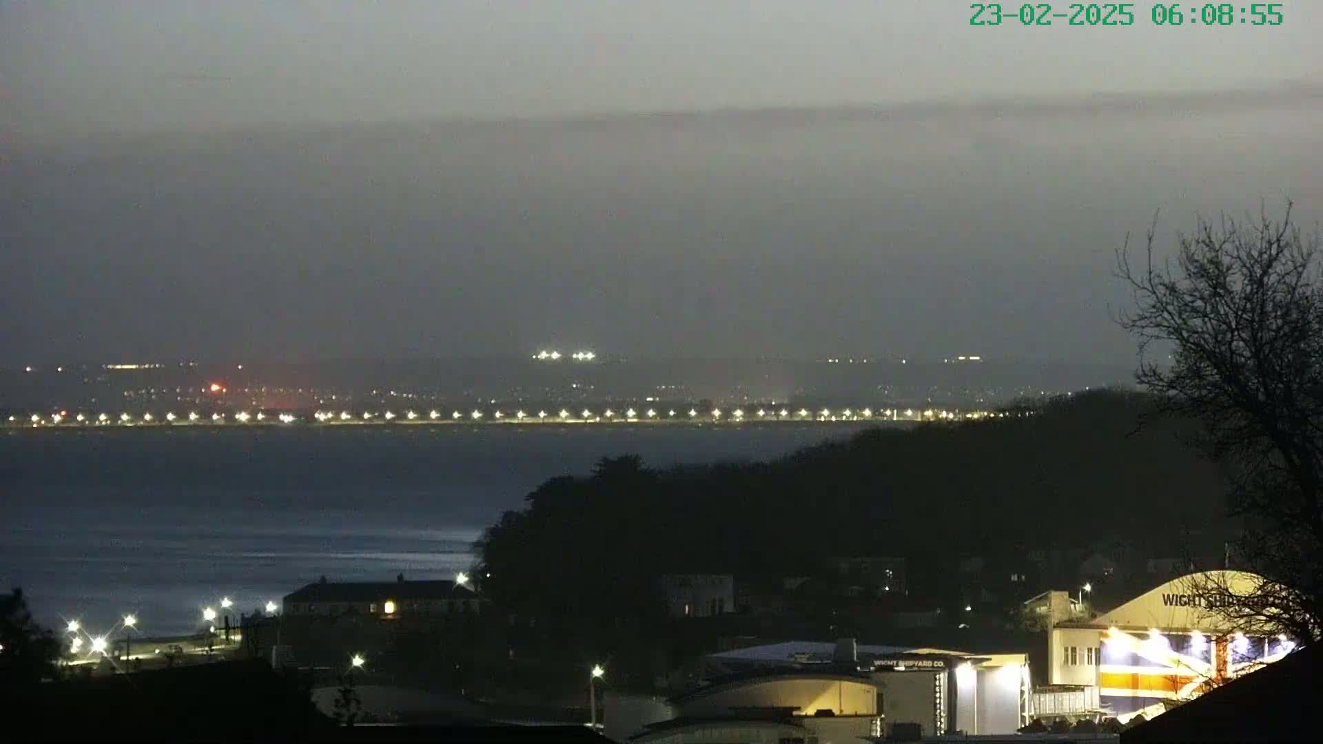 A dimly lit coastal town at night is seen across a dark body of water, with a line of lights visible on the far shore under an overcast sky.