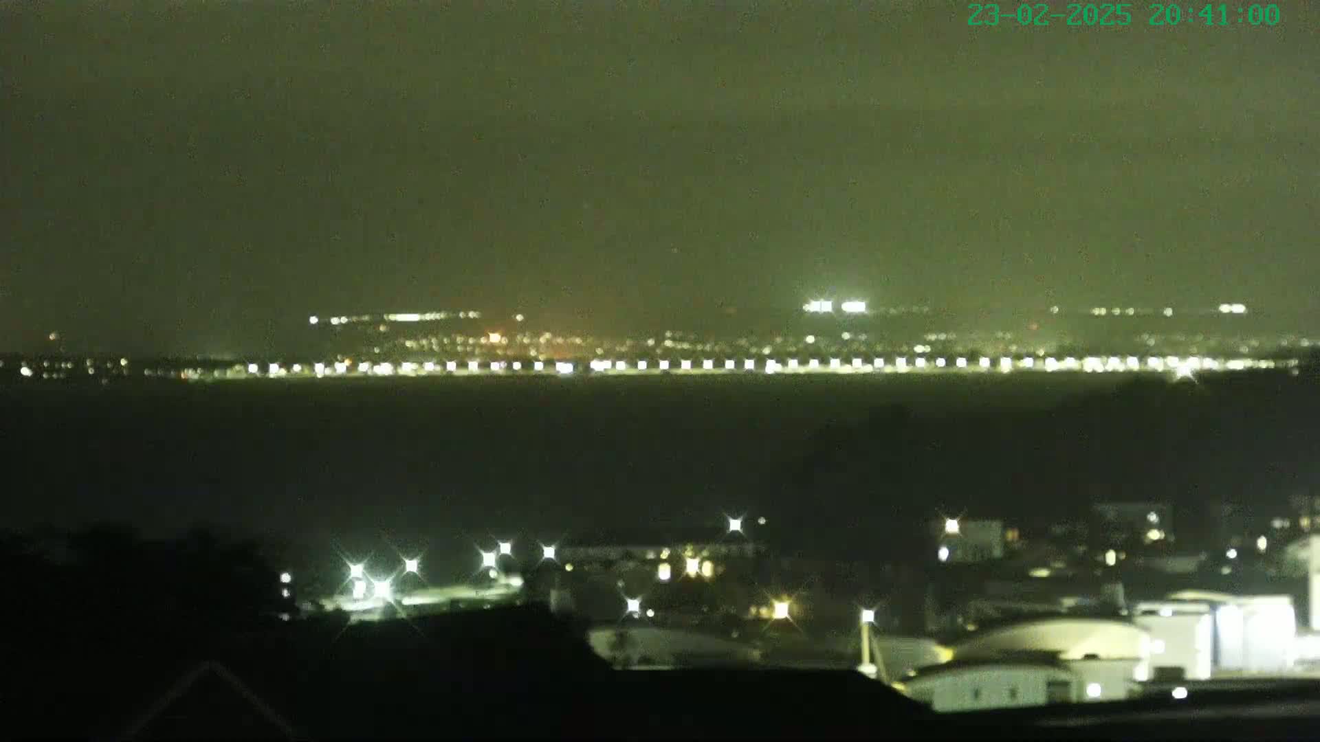 A nighttime view of a city skyline across a body of water, under hazy or overcast conditions.