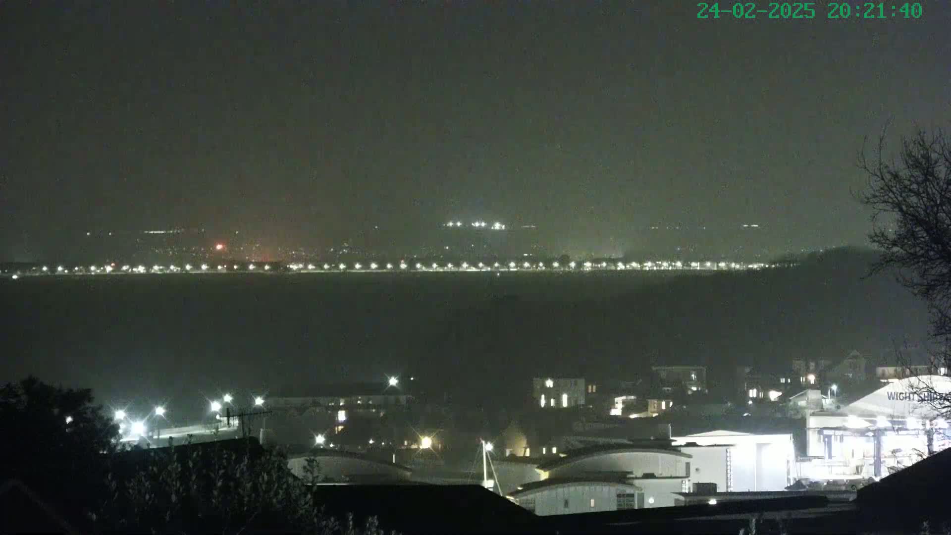 A nighttime view of a hazy town with lights illuminating buildings and a distant shoreline also dotted with lights.